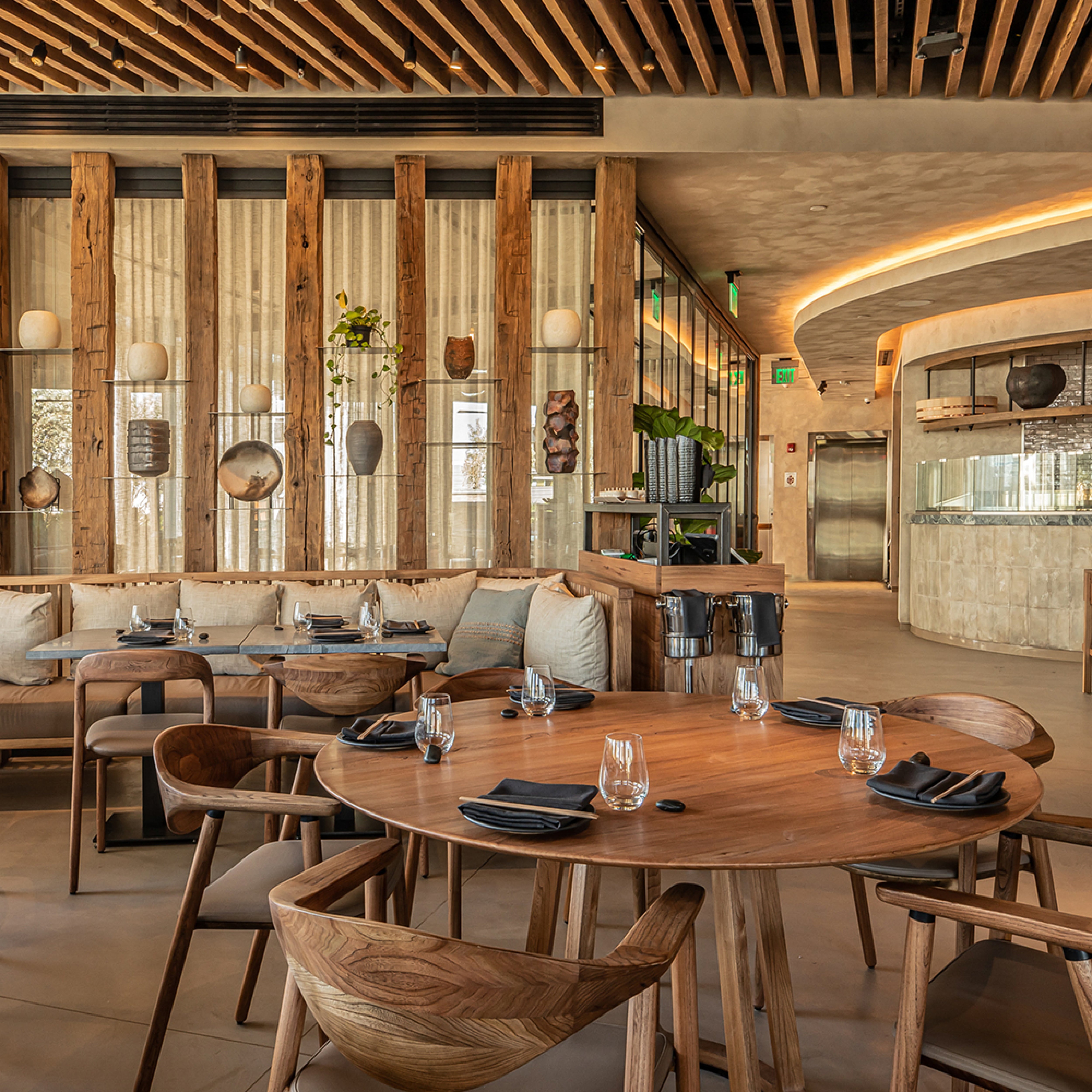 A modern restaurant showcases Artisans NARU Chairs with upholstered seats, solid wood furniture, round tables adorned with glassware and black napkins. Earthy tones, textured walls, and ceiling lights enhance the warm ambiance. An open kitchen area highlights eco-friendly materials in use.