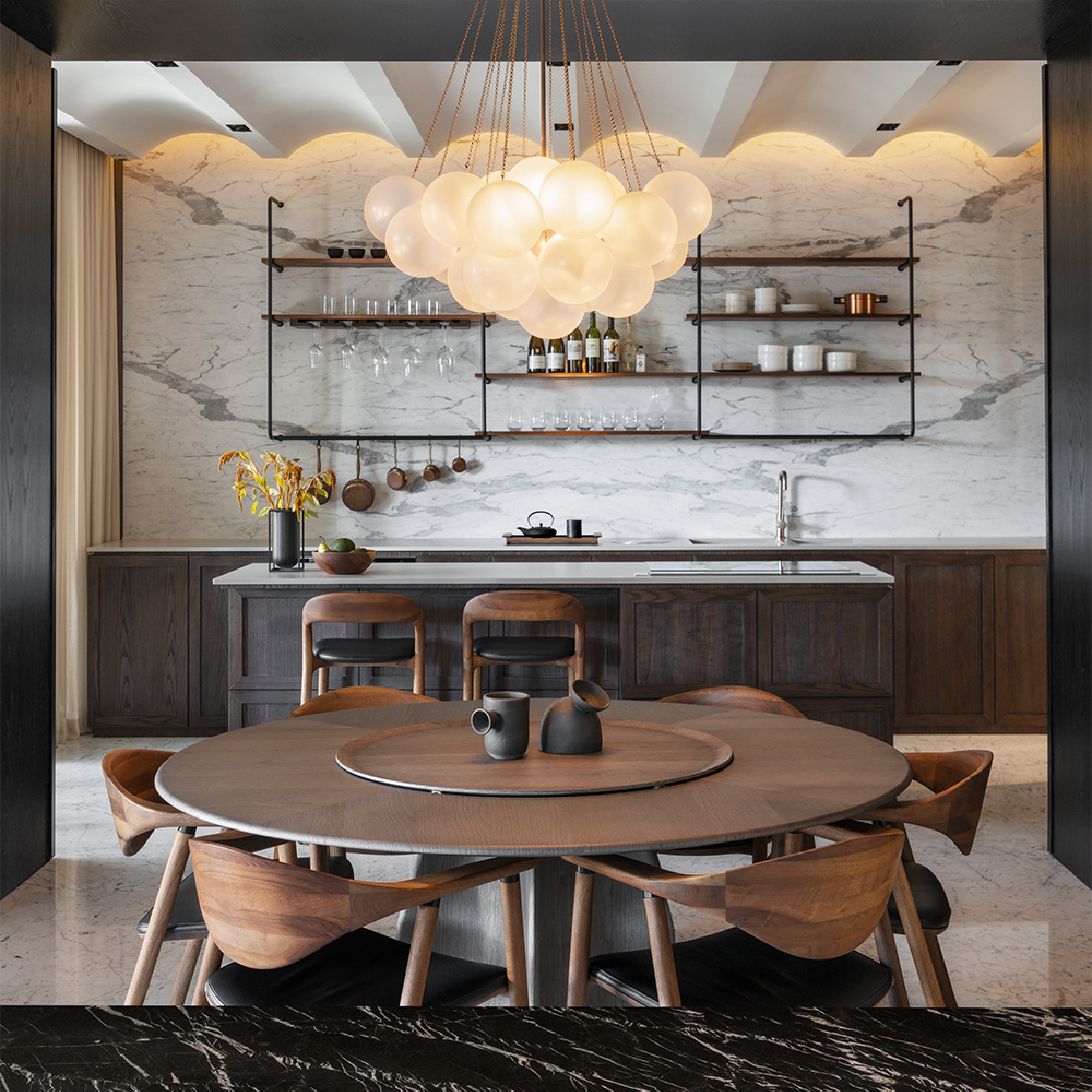 A modern kitchen with a marble backsplash and dark eco-friendly wooden cabinets. A round dining table paired with Artisans NARU Chairs complements the space, where globe pendant lights hang above. Shelves showcase glasses, and warm lighting enhances the inviting atmosphere.
