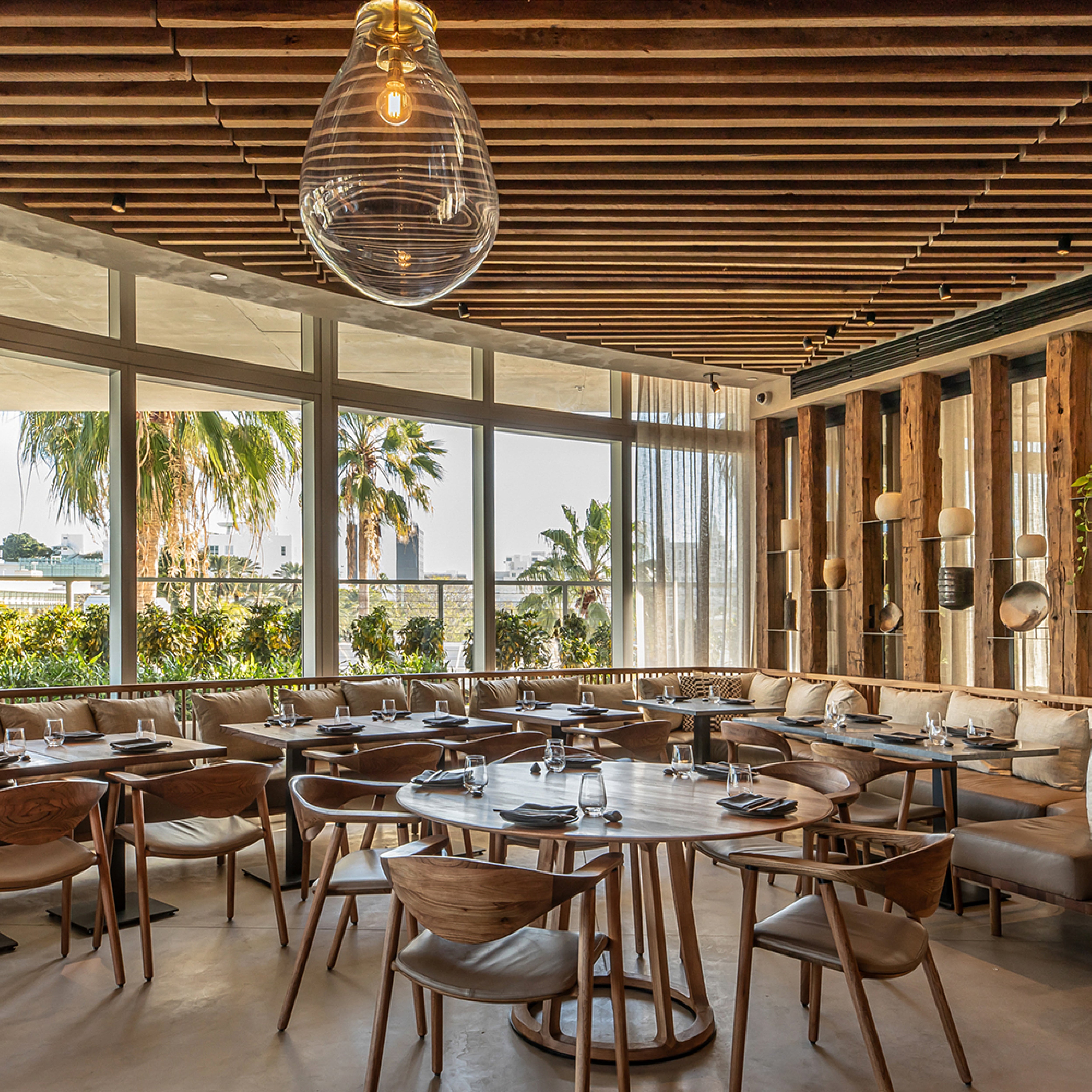 A modern restaurant dining area features round solid wood tables paired with Artisans NARU Chair Upholstered Seat. Large windows reveal palm trees outside, while a wooden slatted ceiling and eco-friendly decorative wall pieces enhance the cozy, elegant ambiance.