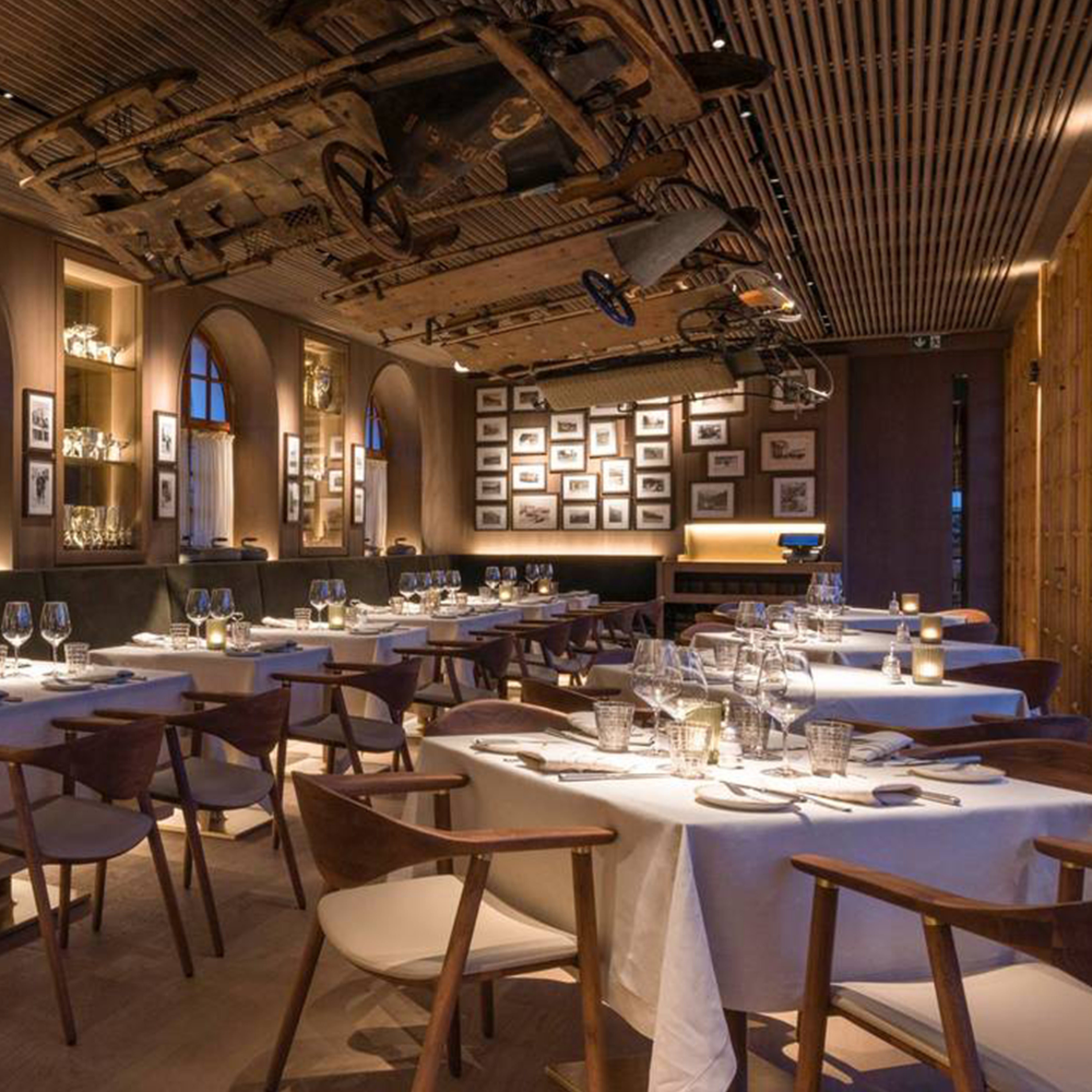 Stylish restaurant interior featuring white tablecloths and Artisans NARU Chair with upholstered seats. The walls are adorned with framed photos, and an antique wooden bicycle made from sustainable materials hangs upside-down on the ceiling. Candlelit tables provide a warm and welcoming atmosphere.