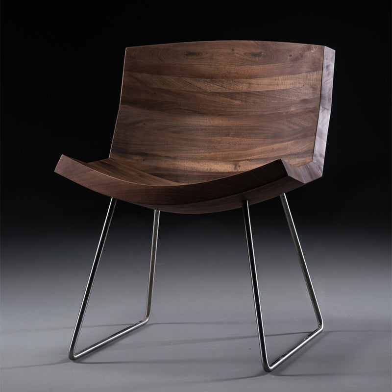 The CHUNK Chair by Artisan features a modern, curved solid wood seat with a rich grain pattern enhanced by natural oils. It sits atop sleek, angular metal legs against a dark gradient background.