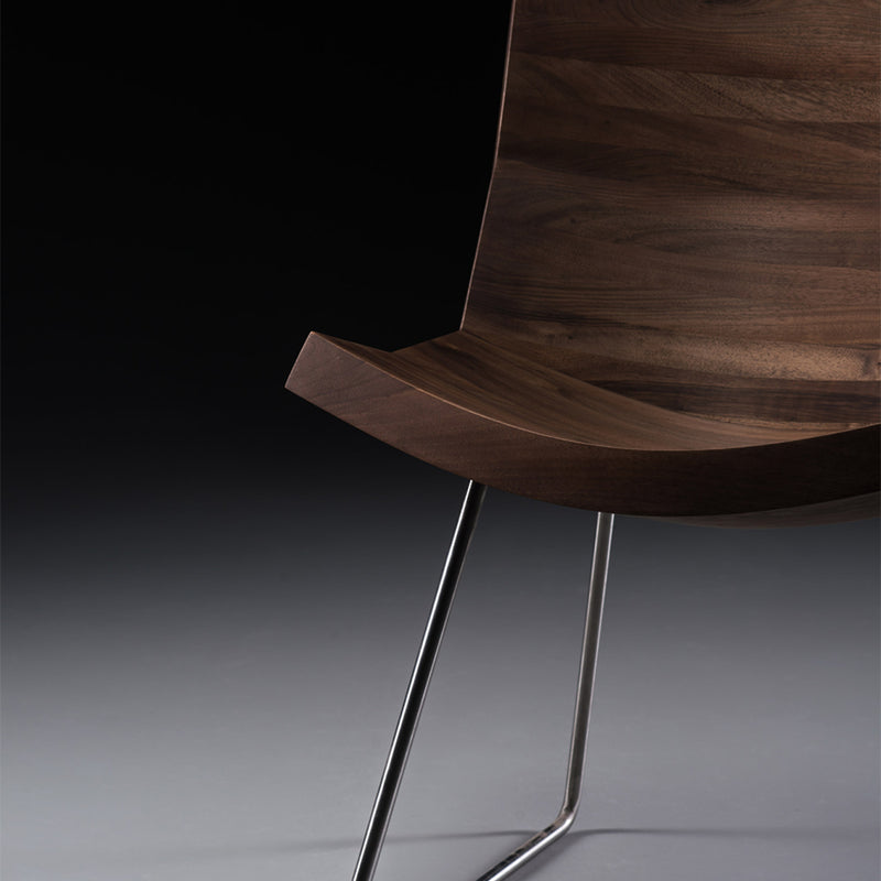 The CHUNK Chair by Artisan features a smooth, dark wooden seat made from solid wood and treated with natural oils. Its curved design and sleek metal legs stand out against the dark background, showcasing its minimalist elegance.