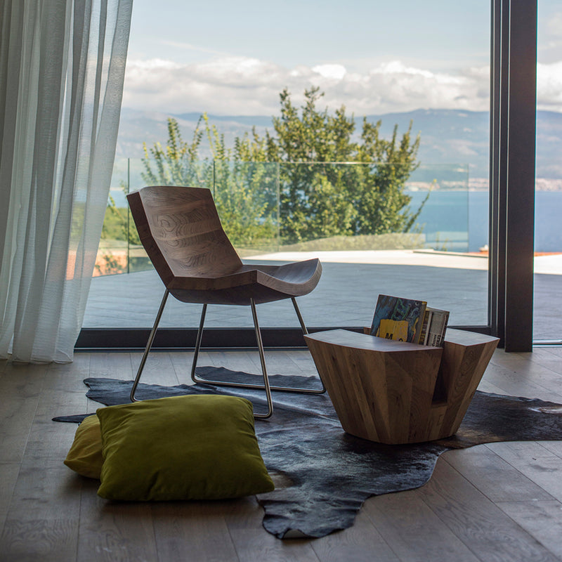 A modern room includes Artisans CHUNK Chair and a geometric table with magazines. Two green cushions rest on a dark rug. Large windows reveal mountains, a lake, and a cloudy sky framed by white curtains, enhanced by the natural oils eco-friendly ambiance.