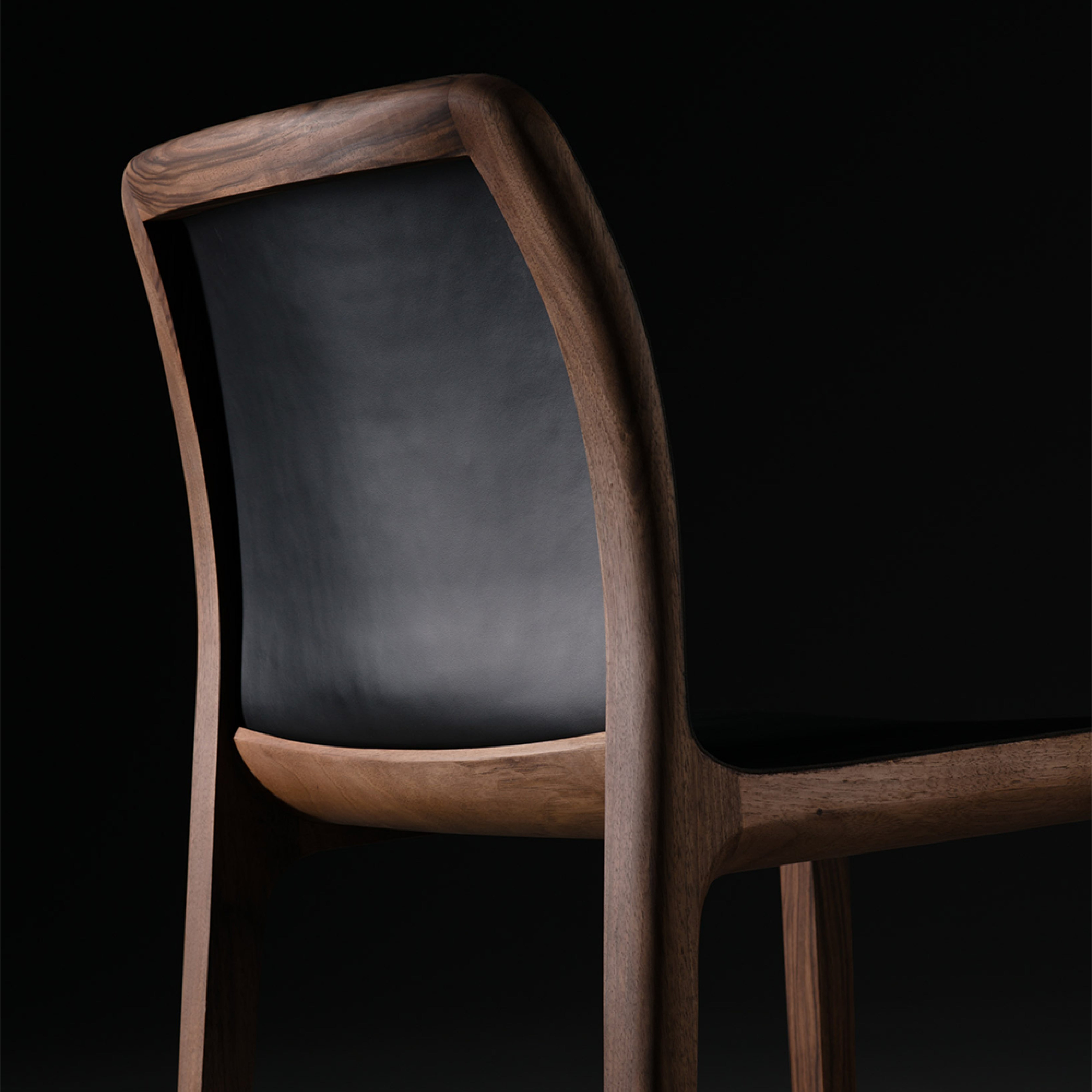 The INVITO Barstool from Artisan, featuring a sleek black leather backrest, stands out against a black background. Its dark solid wood, smooth curved frame, and minimalist design exude elegance and modernity.