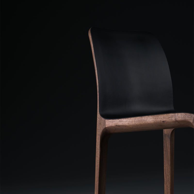 The INVITO Barstool by Artisan is a minimalist wooden chair with a sleek black seat, set against a dark background. Its modern design highlights a curved backrest and solid wood finish.