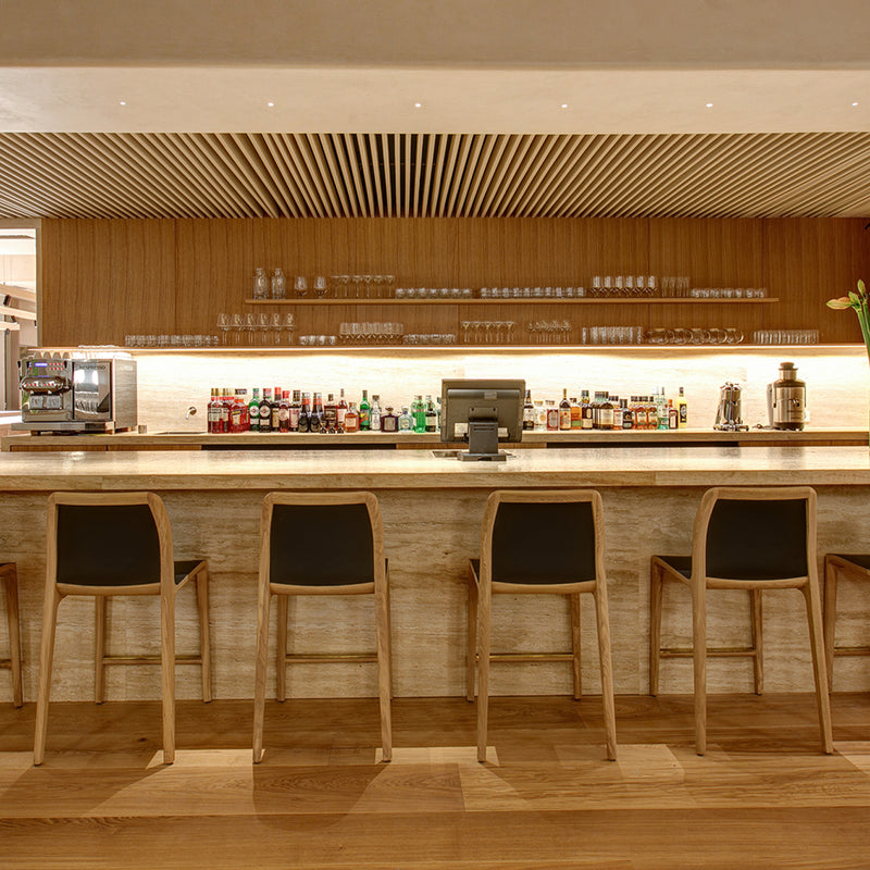 A modern bar features five INVITO Barstools from Artisan, a solid wood finish, a countertop with bottles and mixers, a coffee machine, neatly arranged glasses on wall-mounted shelves, and soft warm lighting to create an inviting atmosphere.