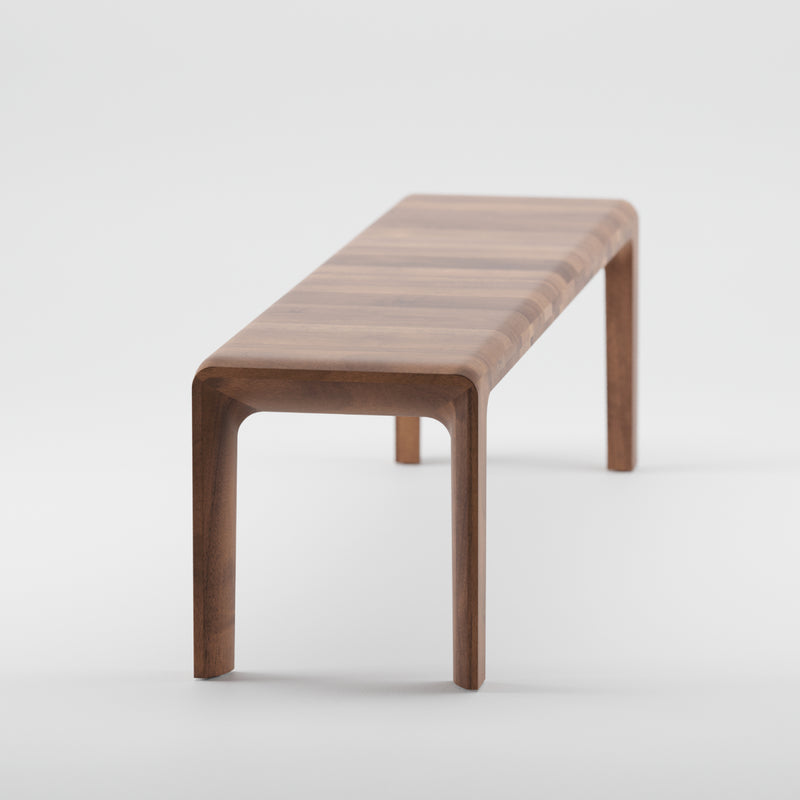The Artisan INVITO Bench features a minimalist design with a smooth solid wood finish. Its simple rectangular seat and four legs are enhanced by natural oils, adding texture and warmth, all set against a plain white background.