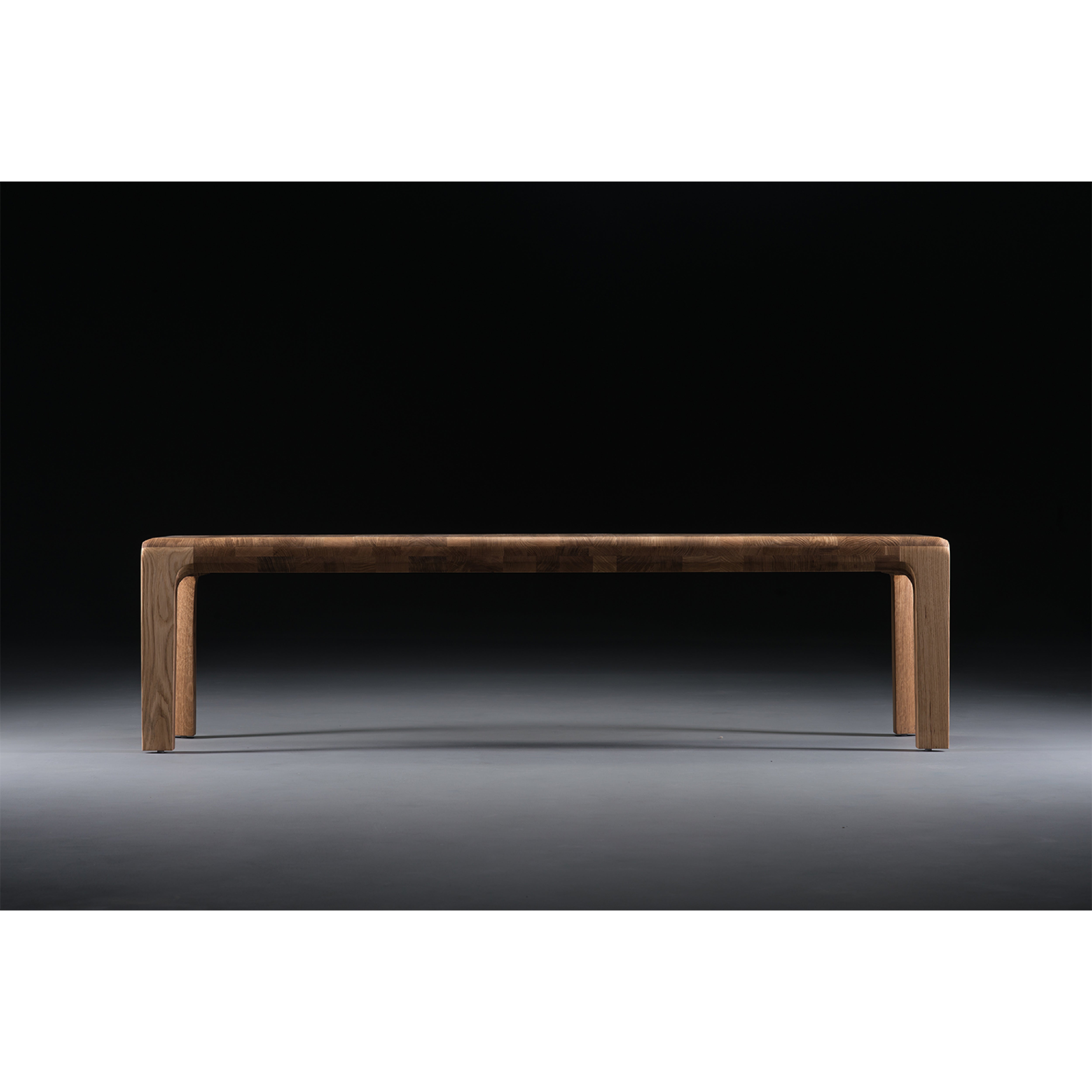 The INVITO Bench by Artisan, a minimalist piece crafted from solid wood and treated with natural oils, stands against a black background, elegantly lit from above to emphasize its sleek surface and clean lines.