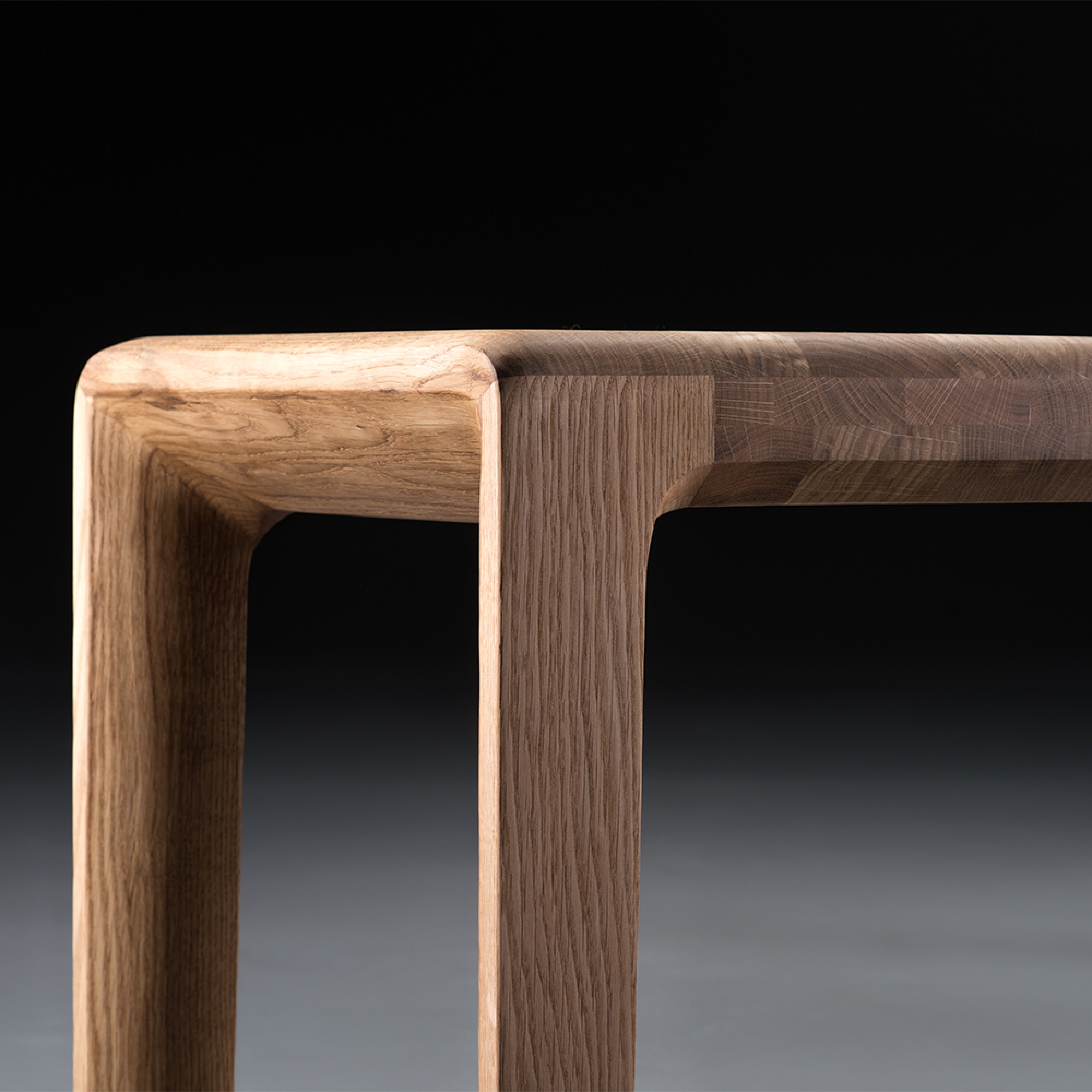 A close-up of the INVITO Bench by Artisan reveals a wooden table corner crafted from solid wood with a smooth, light-brown finish enhanced by natural oils. The tabletop and legs are seamlessly connected with clean, modern lines against a dark gradient background.