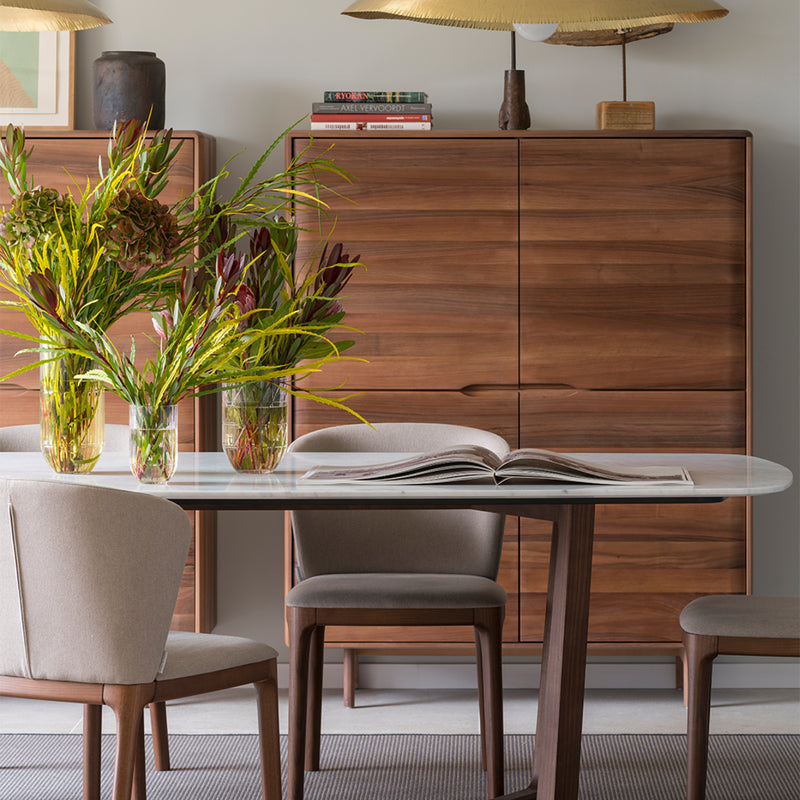 A modern dining room features a solid wood table set for four. Three vases with green and brown flowers sit next to an open book. The Artisan INVITO Highboard showcases books and eco-friendly decor beneath a large, golden pendant light.