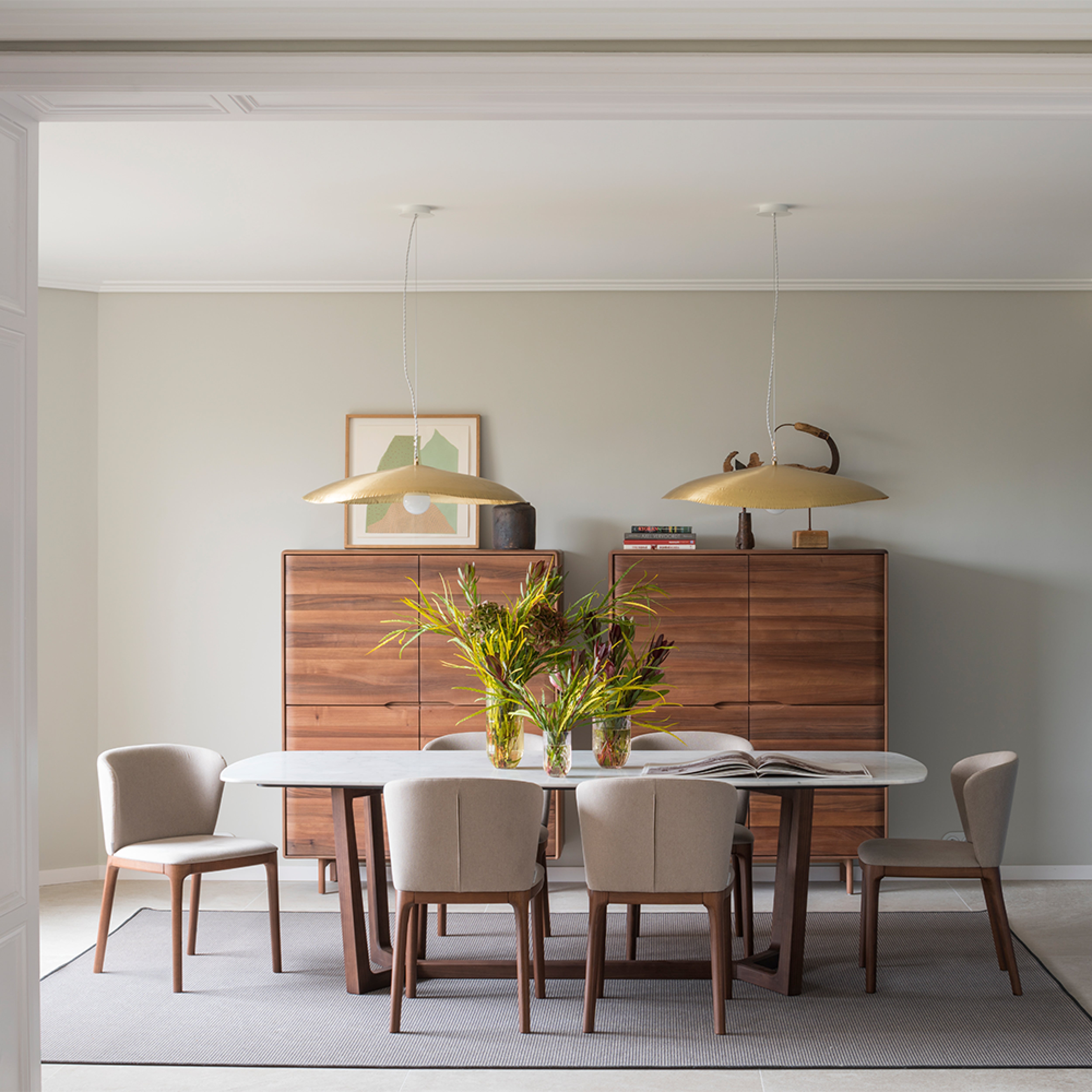 In a modern dining room, a white oval table with six beige chairs is illuminated by two pendant lights overhead. The Artisan INVITO Highboard showcases art and decorations in the background. A vase of green and yellow flowers, enhanced by natural oils, sits at the center of the table.