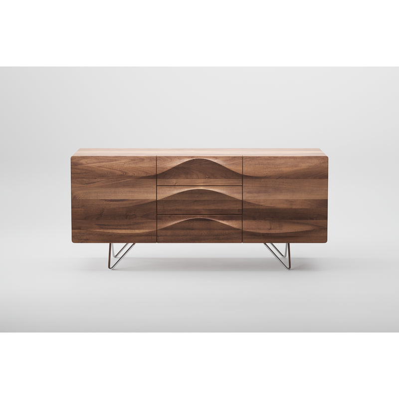The LASTA Sideboard by Artisan is a modern, environmentally-friendly piece crafted with natural oils. It features a wave pattern, three drawers, and two cabinet doors. Elevated on minimalist metal legs, this solid wood sideboard is set against a plain white background.