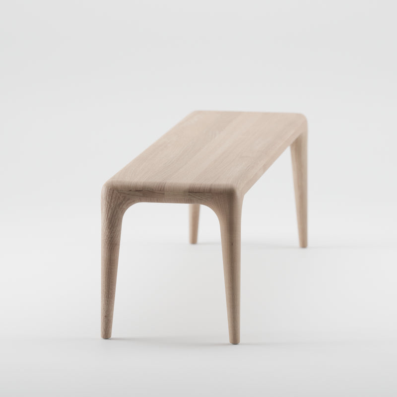 The LATUS Bench by Artisan is a minimalist wooden bench crafted from solid wood, featuring sleek legs and a smooth, light finish enhanced by natural oils, all set against a white background.