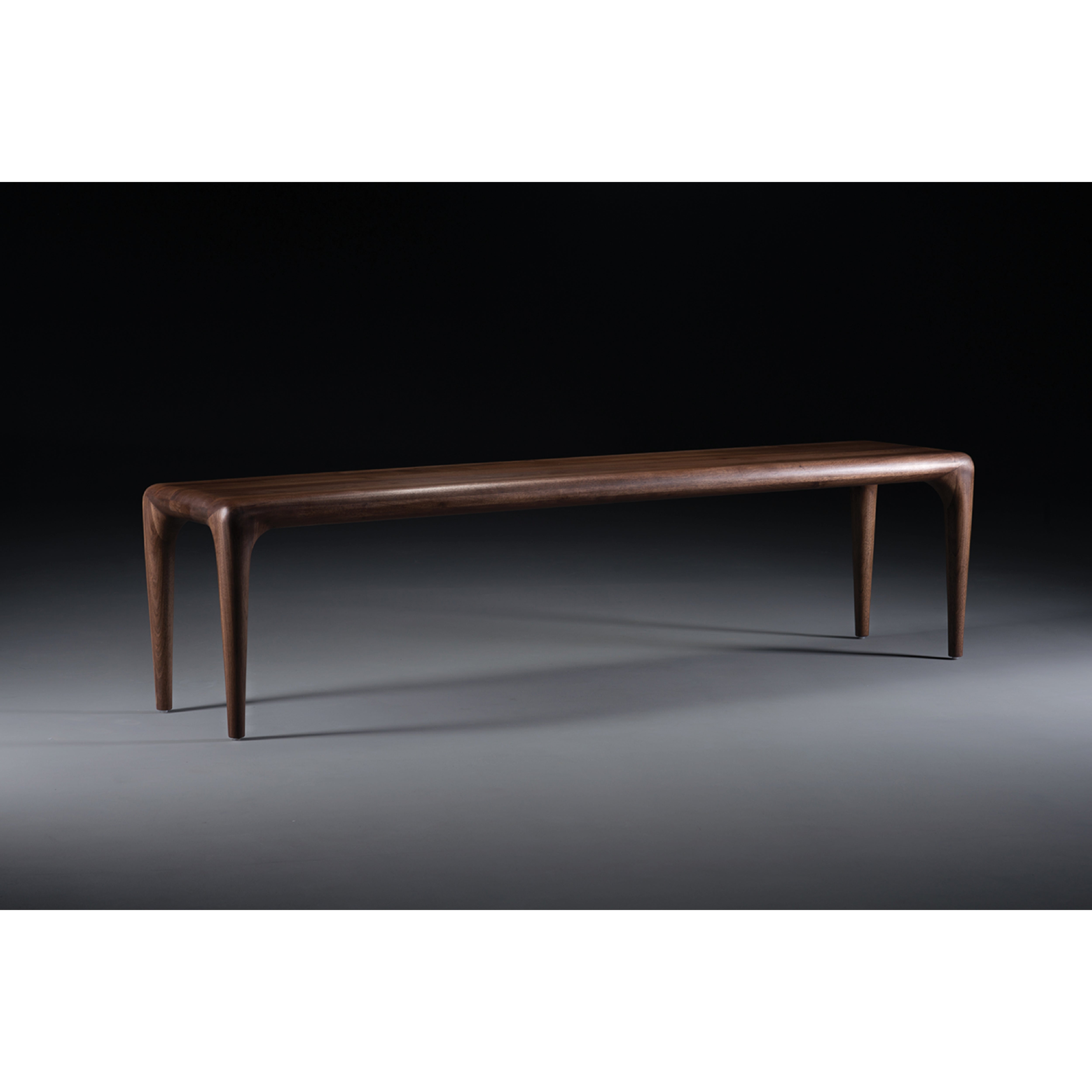 The LATUS Bench by Artisan is a sleek wooden bench crafted from solid wood, featuring smooth, rounded edges. Enhanced with natural oils, it has four slender, tapered legs and a polished surface that exudes minimalist and modern charm against a dark background.