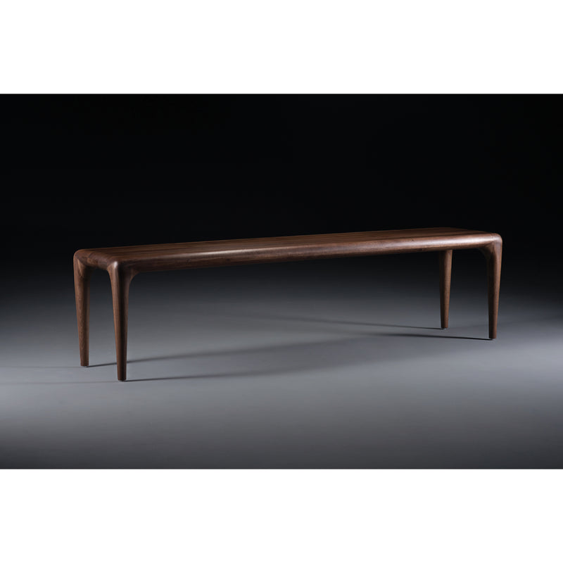 The LATUS Bench by Artisan is a sleek wooden bench crafted from solid wood, featuring smooth, rounded edges. Enhanced with natural oils, it has four slender, tapered legs and a polished surface that exudes minimalist and modern charm against a dark background.
