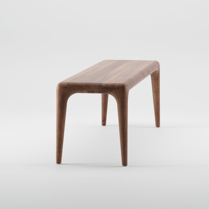 The LATUS Bench by Artisan is a sleek wooden piece crafted from solid wood with a smooth finish and rounded corners. Enhanced by natural oils, it features four tapered legs and a minimalist design, shown against a plain white background.