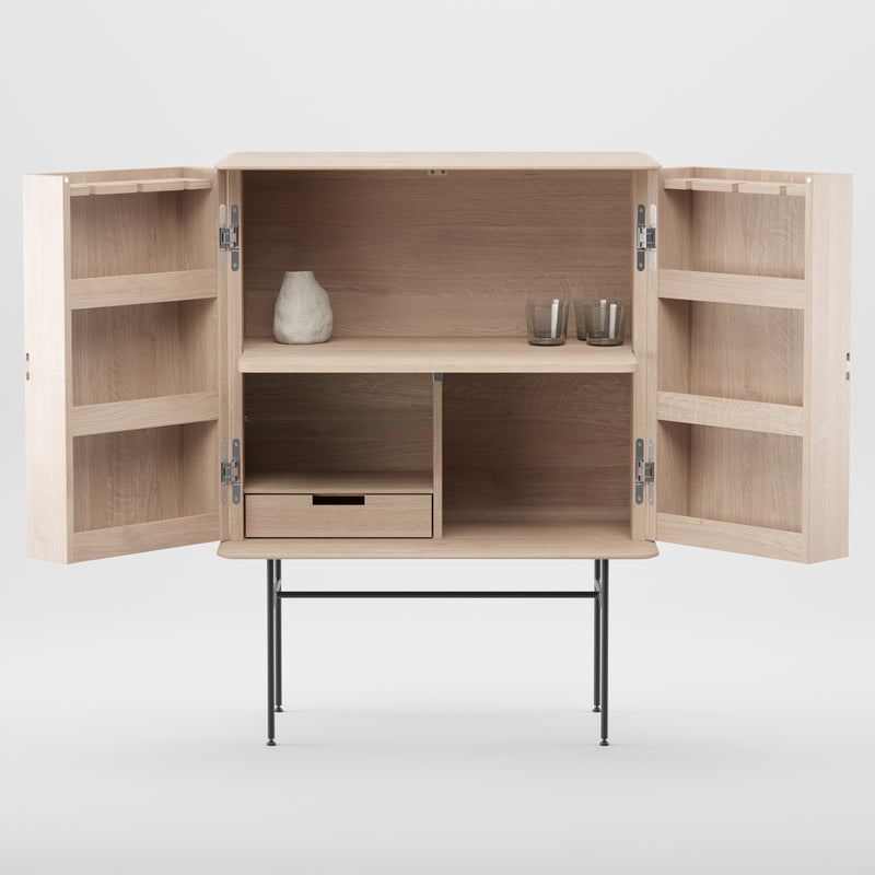 The Leno Bar by Artisan features modern design with light wood, open doors revealing minimalistic shelving adorned with a vase and two glasses. An open shelf complements a closed drawer below. This artisan furniture rests on slim black metal legs.