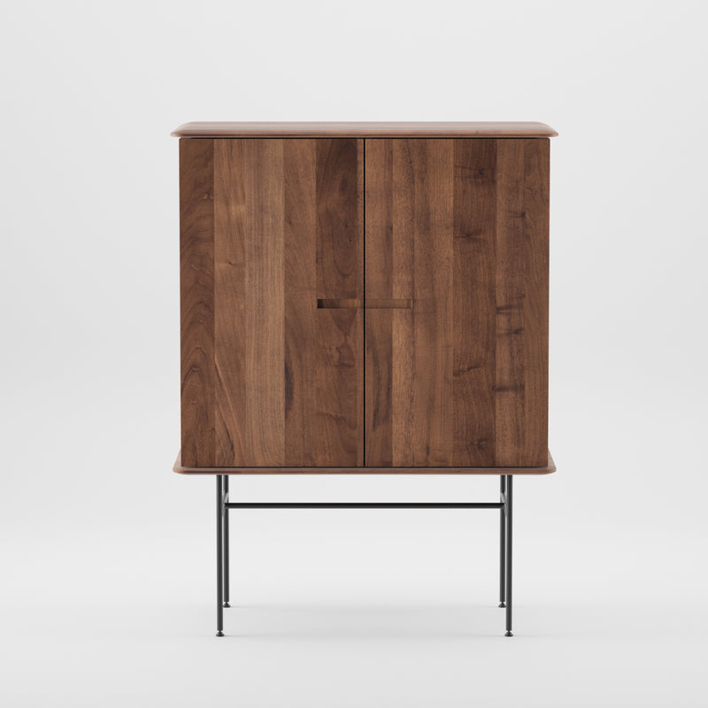The Artisan Leno Bar is a wooden cabinet with a smooth dark finish and slim metal legs. Expertly crafted, it has two doors with seamless handles for a modern, sleek look against a white background.