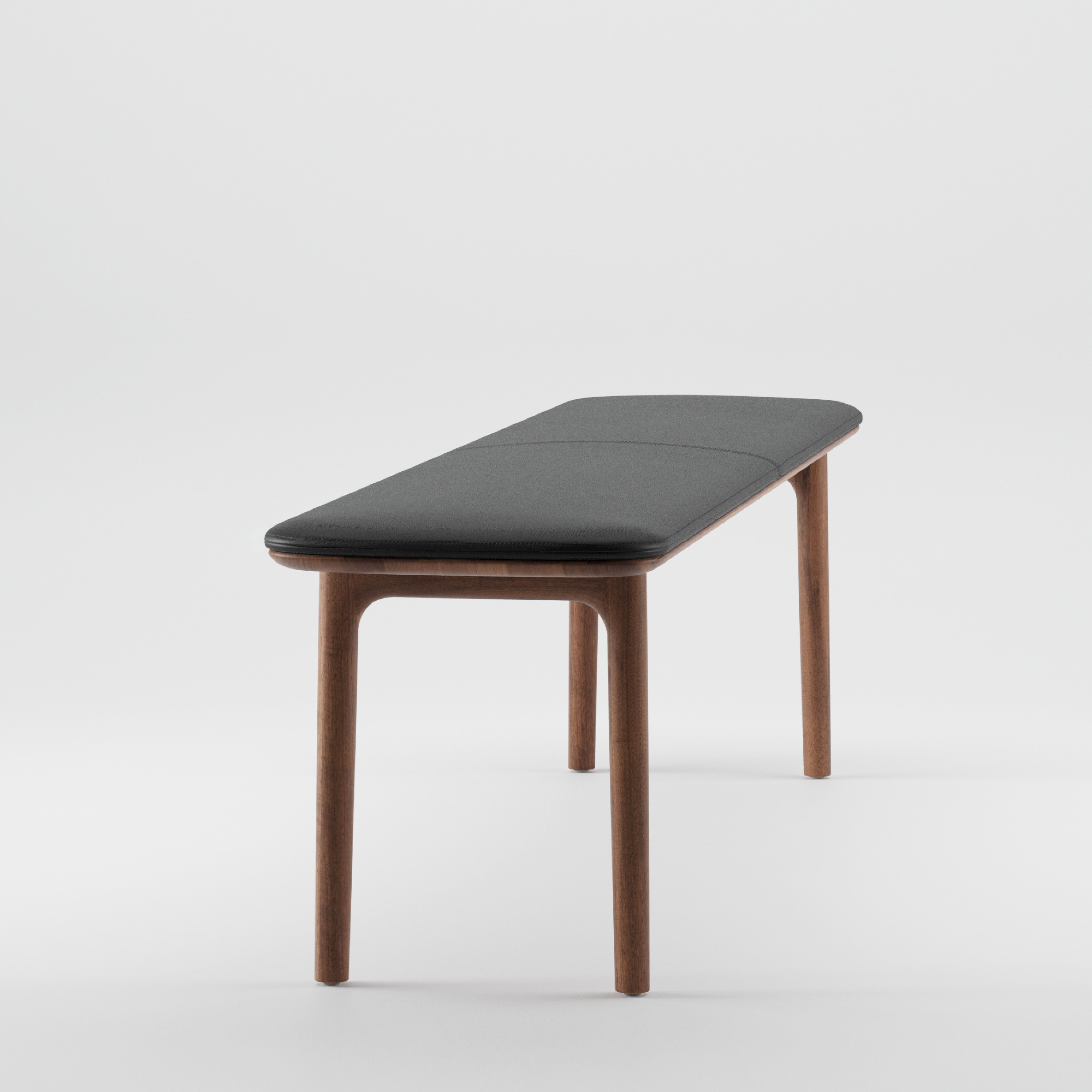 The NEVA Bench by Artisan, showcasing a dark cushioned seat and four wooden legs, is presented against a plain white background. Its minimalist design highlights simplicity and functionality with organic wood forms.