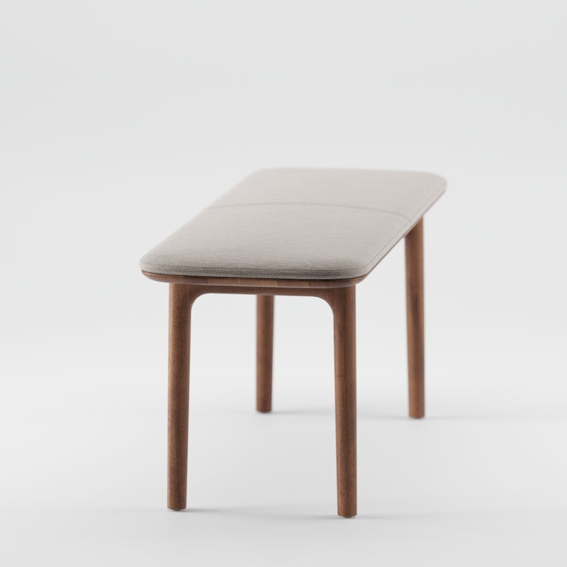 The NEVA Bench by Artisan is a minimalist piece featuring a light gray cushioned seat and four slender legs. Part of the Neva collection, this bench combines modern simplicity with organic wood forms against a white backdrop.