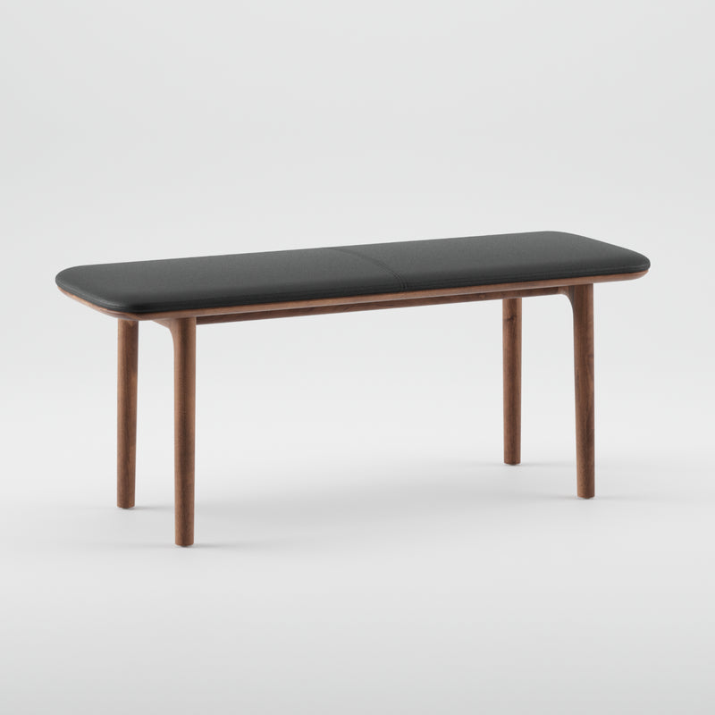 The NEVA Bench by Artisan showcases a contemporary design with a dark cushioned seat and sleek wooden legs, highlighted against a white background with organic wood forms.
