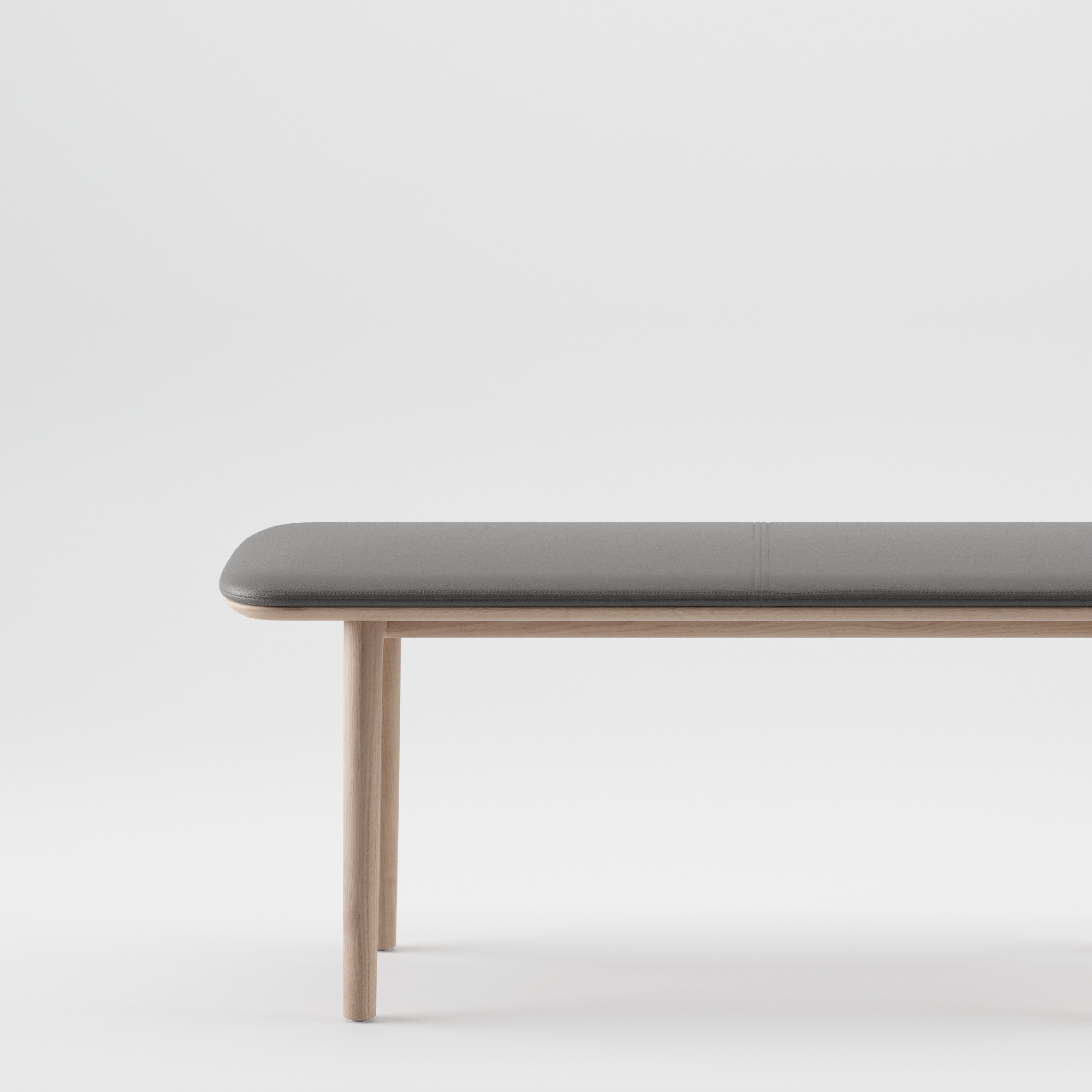 Artisans NEVA Bench boasts a minimalist design with a sleek gray cushioned seat and light wooden legs, highlighting organic wood forms against a plain white background.