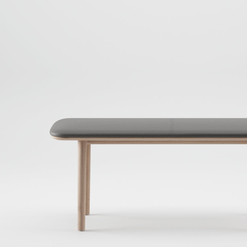 Artisans NEVA Bench boasts a minimalist design with a sleek gray cushioned seat and light wooden legs, highlighting organic wood forms against a plain white background.