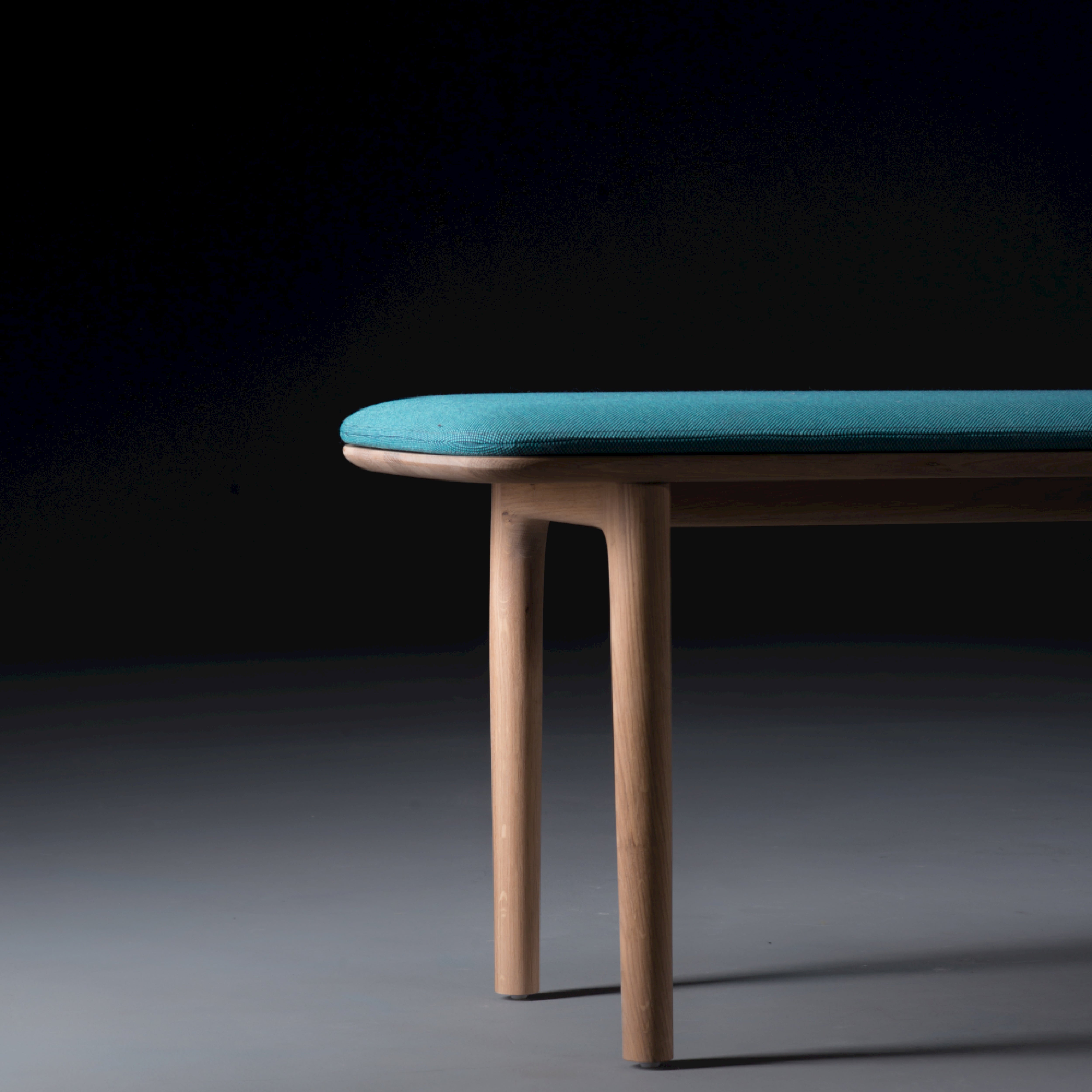The image depicts the NEVA Bench by Artisan, featuring organic wood forms and a teal upholstered seat against a dark background. Viewed from the side, it highlights its textured design and craftsmanship as part of the Neva collection.