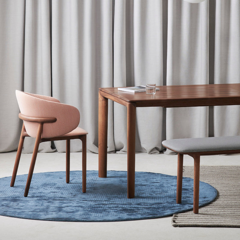 A minimalist dining setup features a wooden table with organic forms, a pink upholstered chair, and the Artisan NEVA Bench with gray cushions. The setup sits on a blue circular rug, framed by light gray curtains in the background.