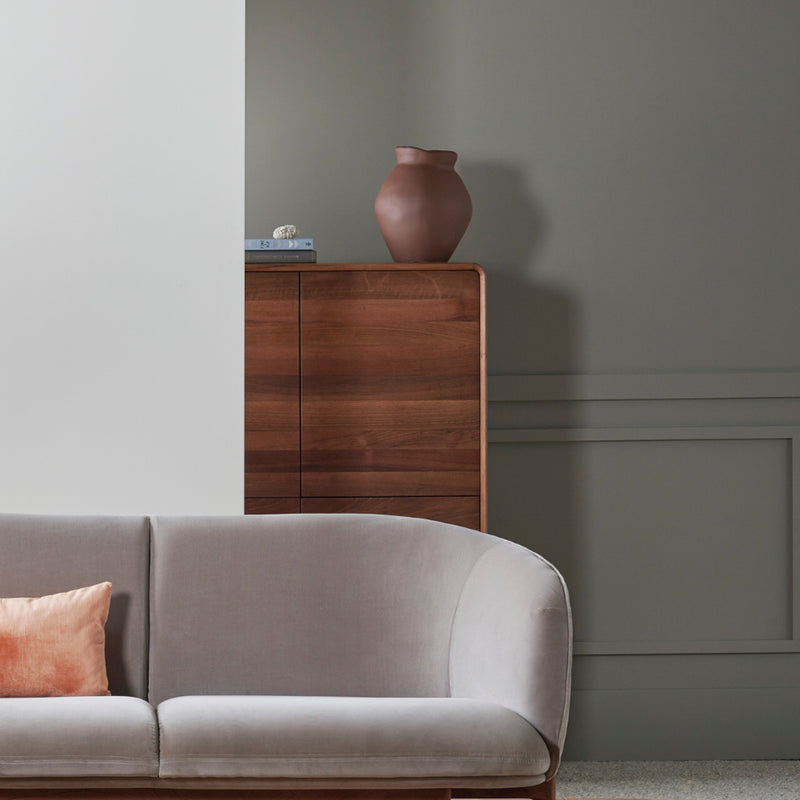 A modern living room showcases a light gray curved sofa with a peach pillow. Against the gray paneled wall, an Artisan NEVA Highboard with natural finishes displays a reddish-brown artisanal vase and stacked books.