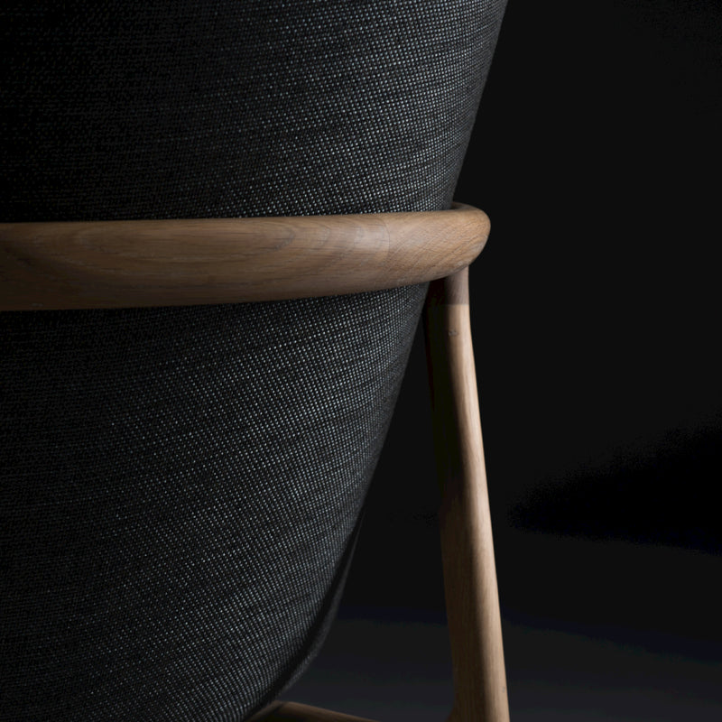Close-up of the Artisan NEVA Lounge Chair High, featuring a textured dark fabric back and smooth wooden frame, combining modern technology with artisanal craftsmanship against a black background.