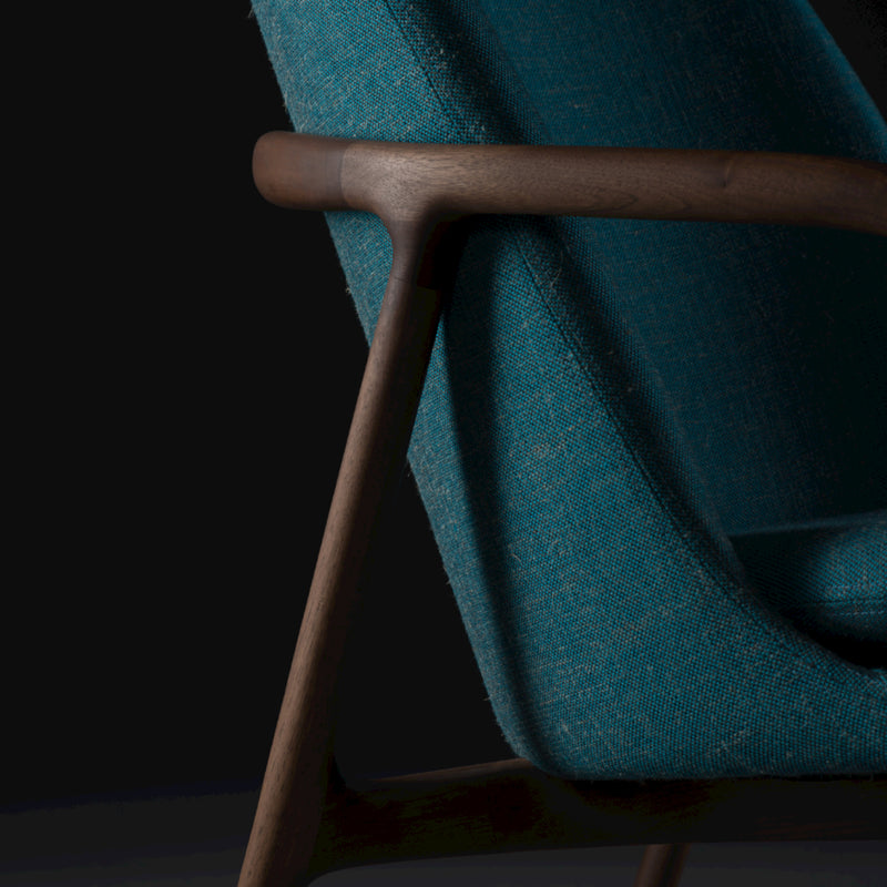 Close-up of the Artisan NEVA Lounge Chair High, featuring a teal fabric seat and sleek wooden frame. The dark background highlights its vibrant color and clean lines, combining modern tech with artisanal craftsmanship for a contemporary touch.