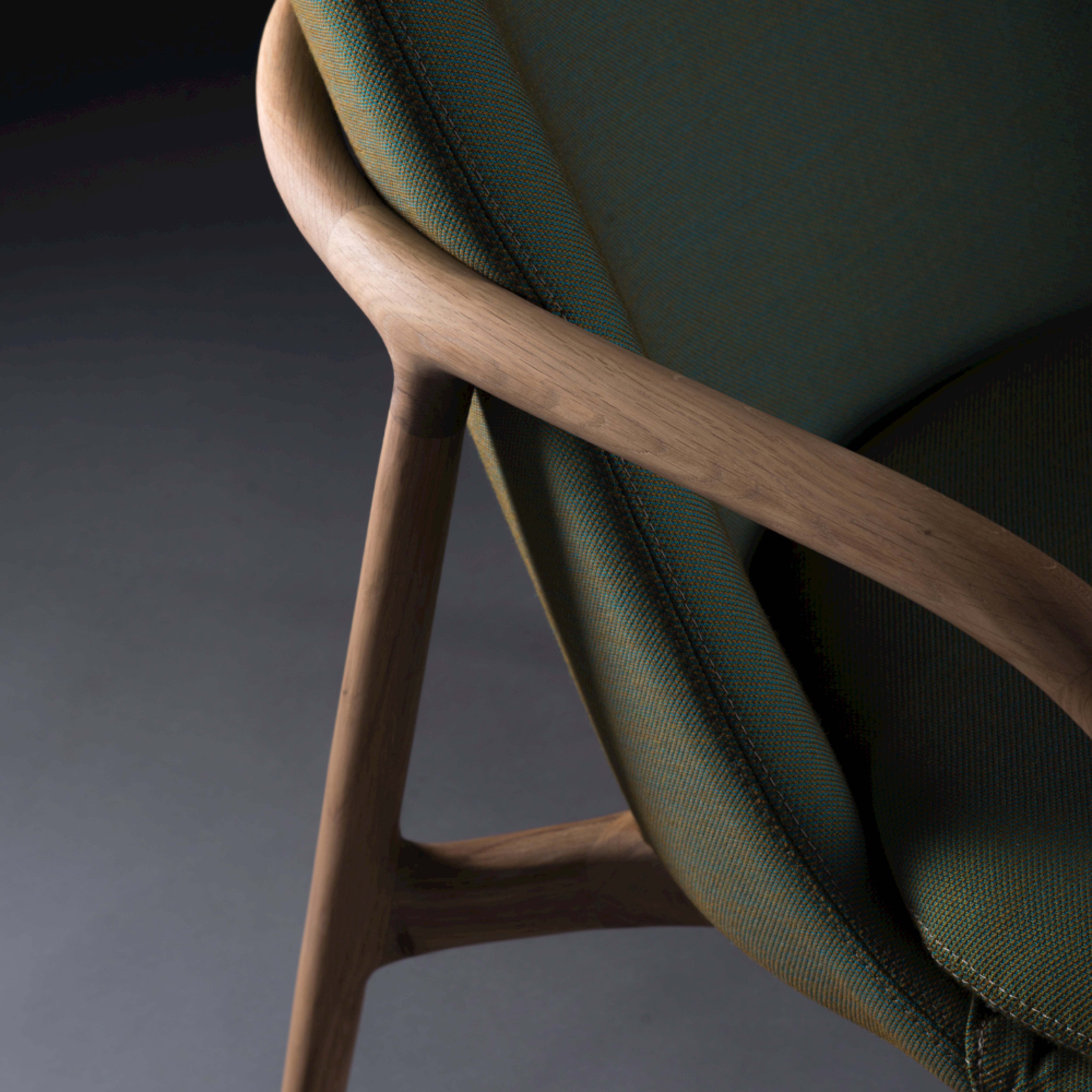 Close-up of an Artisan NEVA Lounge Chair High with a wooden frame and green fabric upholstery showcases the smooth, curved armrest and backrest. A dark background enhances its modern yet artisanal design.