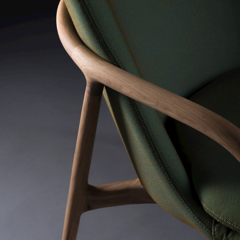 Close-up of an Artisan NEVA Lounge Chair High with a wooden frame and green fabric upholstery showcases the smooth, curved armrest and backrest. A dark background enhances its modern yet artisanal design.
