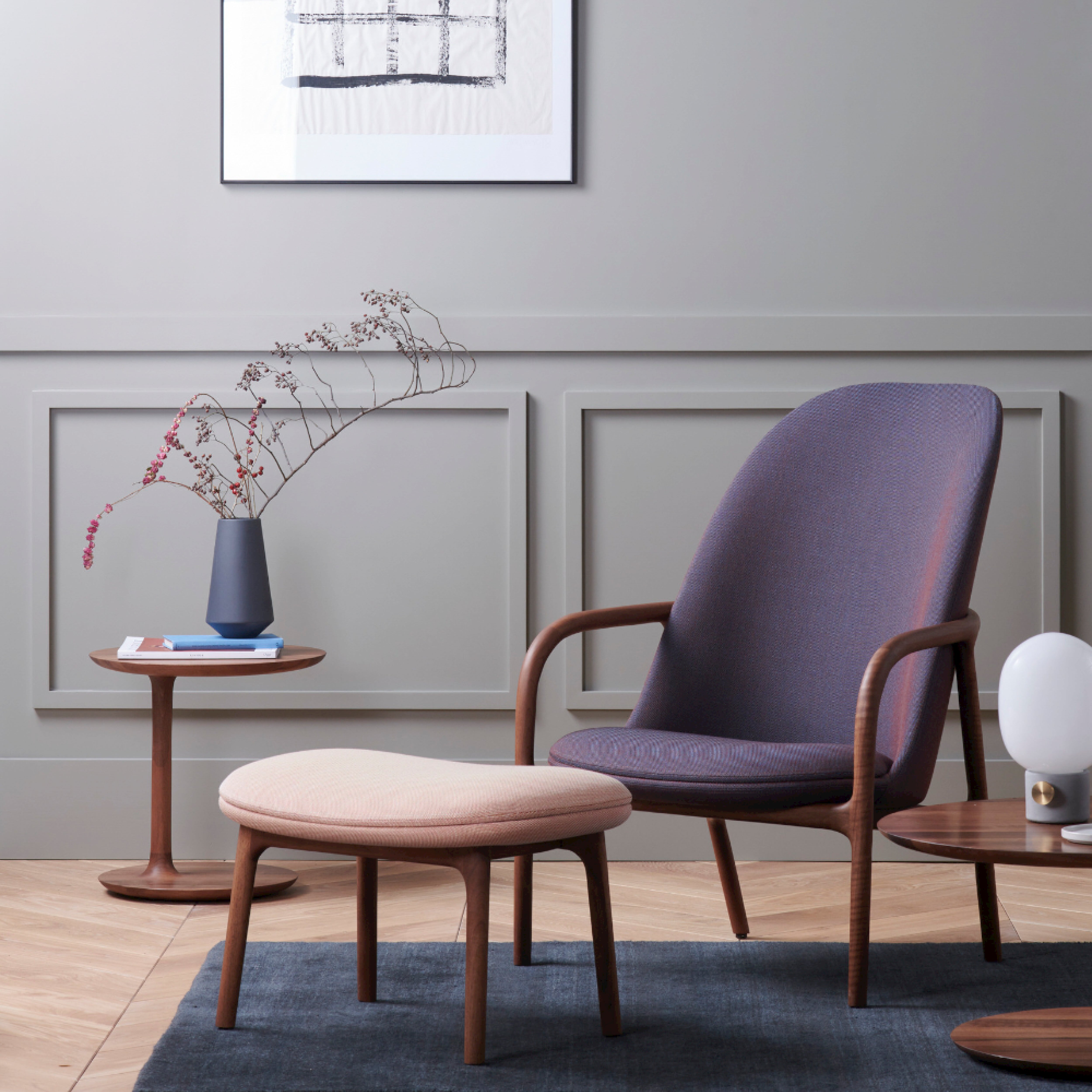 A modern living room features a NEVA Lounge Chair High by Artisan with a matching ottoman on a blue rug. Two round wooden tables hold a vase with branches and a lamp. A geometric framed artwork adorns the light gray paneled wall, merging modern technology with artisanal craftsmanship seamlessly.