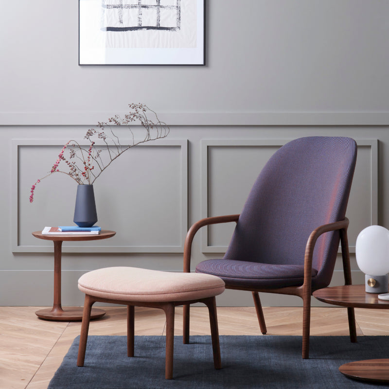 A modern living room features a NEVA Lounge Chair High by Artisan with a matching ottoman on a blue rug. Two round wooden tables hold a vase with branches and a lamp. A geometric framed artwork adorns the light gray paneled wall, merging modern technology with artisanal craftsmanship seamlessly.
