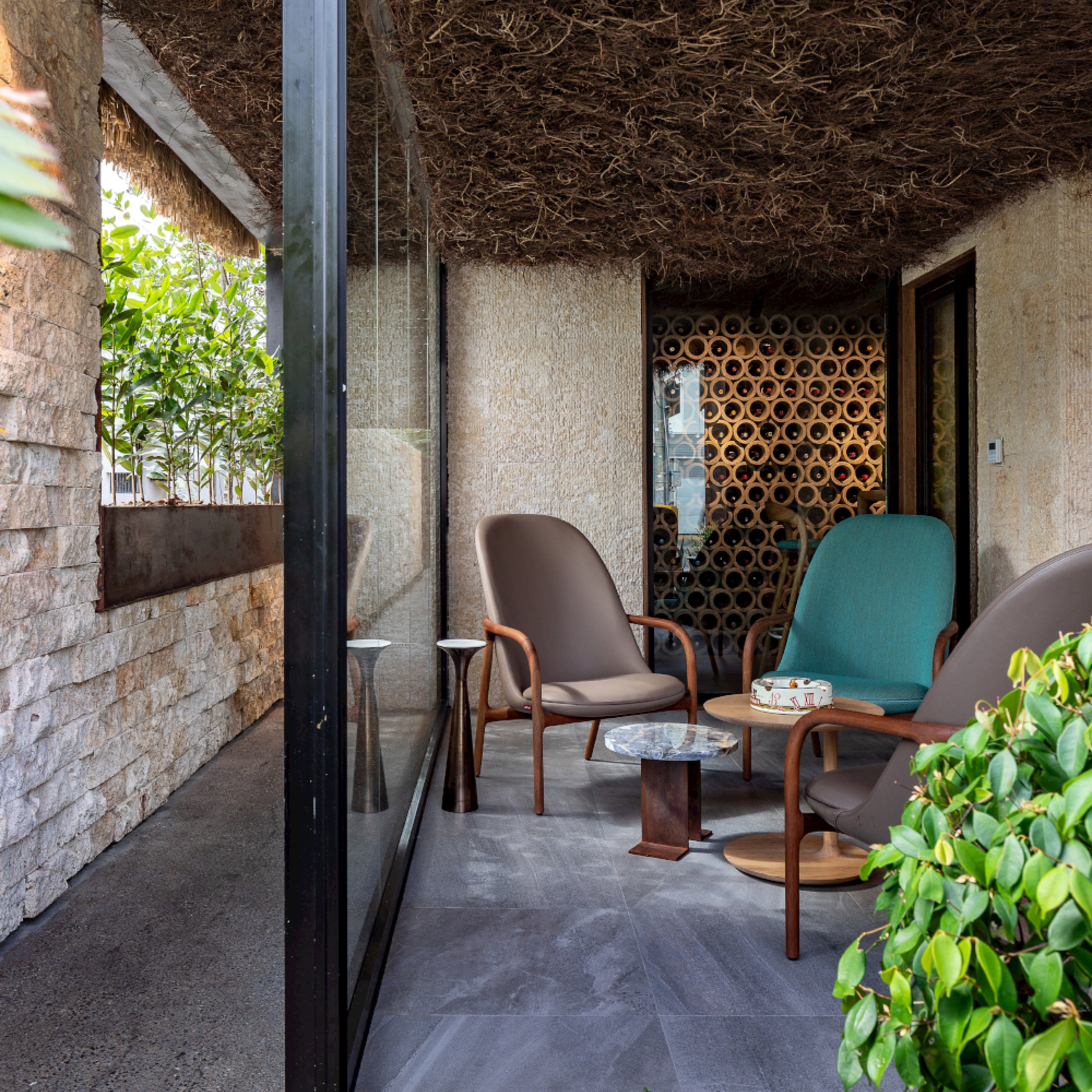 Four Artisan NEVA Lounge Chair Highs form a cozy outdoor seating area around small tables. A natural, textured ceiling and walls with plants on each side enhance the ambiance. The slate grey floor complements the space, while a glass partition integrates modern technology with nature.