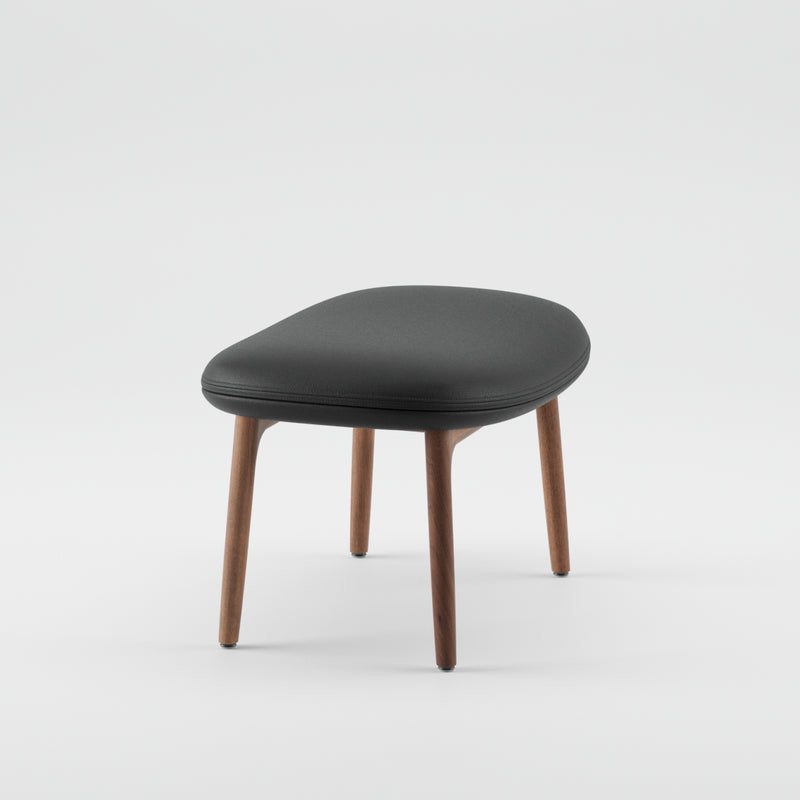 The NEVA Ottoman by Artisan epitomizes Scandinavian lounge furniture with its sleek black cushioned seat and four angled wooden legs, set against a plain white background.