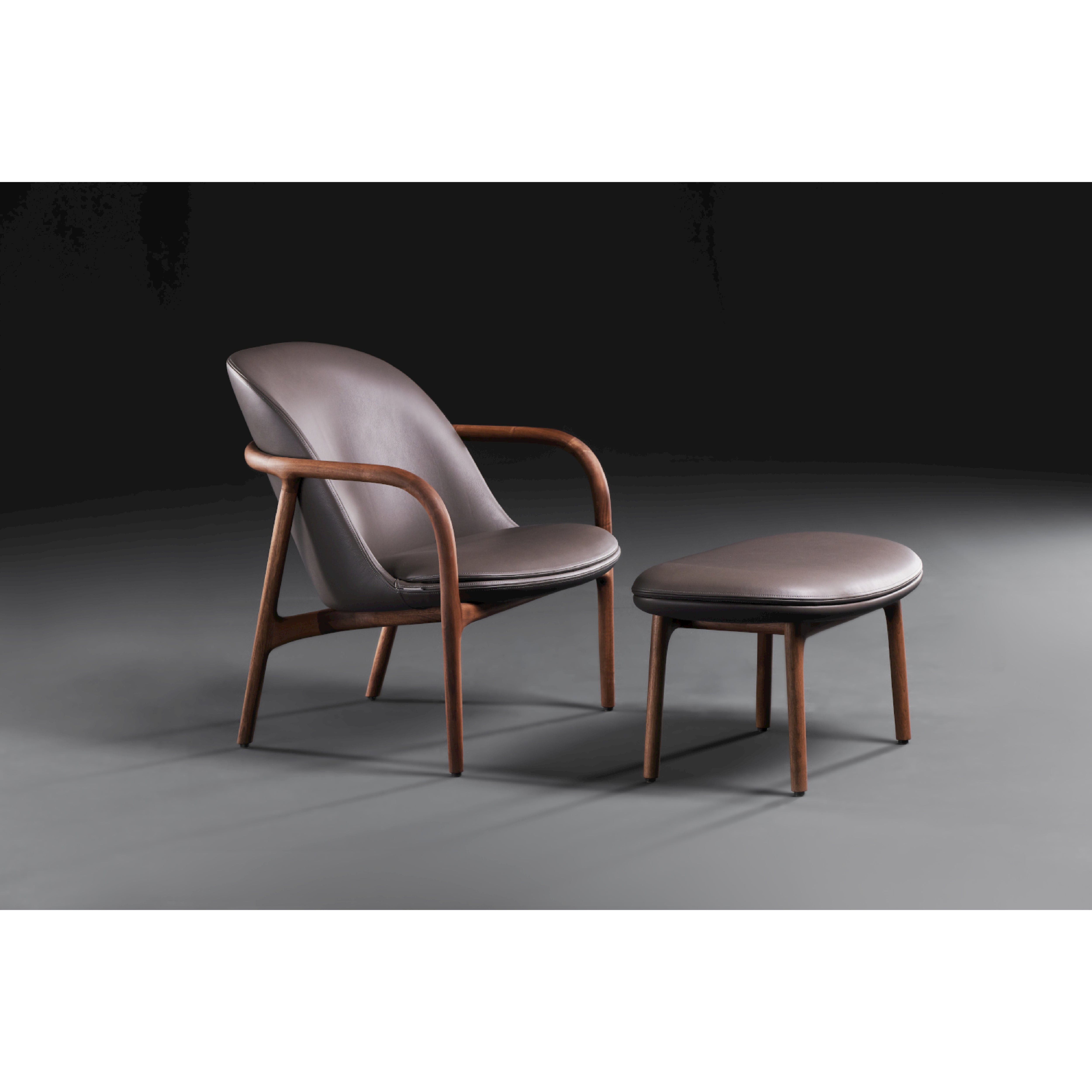 The NEVA Ottoman by Artisan is a Scandinavian lounge chair and ottoman set with a sleek dark wooden frame and brown leather upholstery. It features a curved back and armrests, resting on a smooth gray surface against a dark background, highlighting exquisite artisanal craftsmanship.