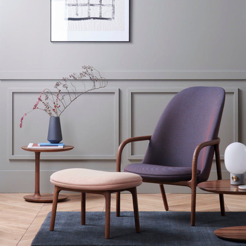 A contemporary living room corner highlights an Artisan NEVA Ottoman with a cushioned chair on a dark rug, embodying modern Scandinavian lounge furniture. A side table holds a vase with branches, another supports a lamp, and abstract art hangs against wainscoting, enhancing the inviting atmosphere.