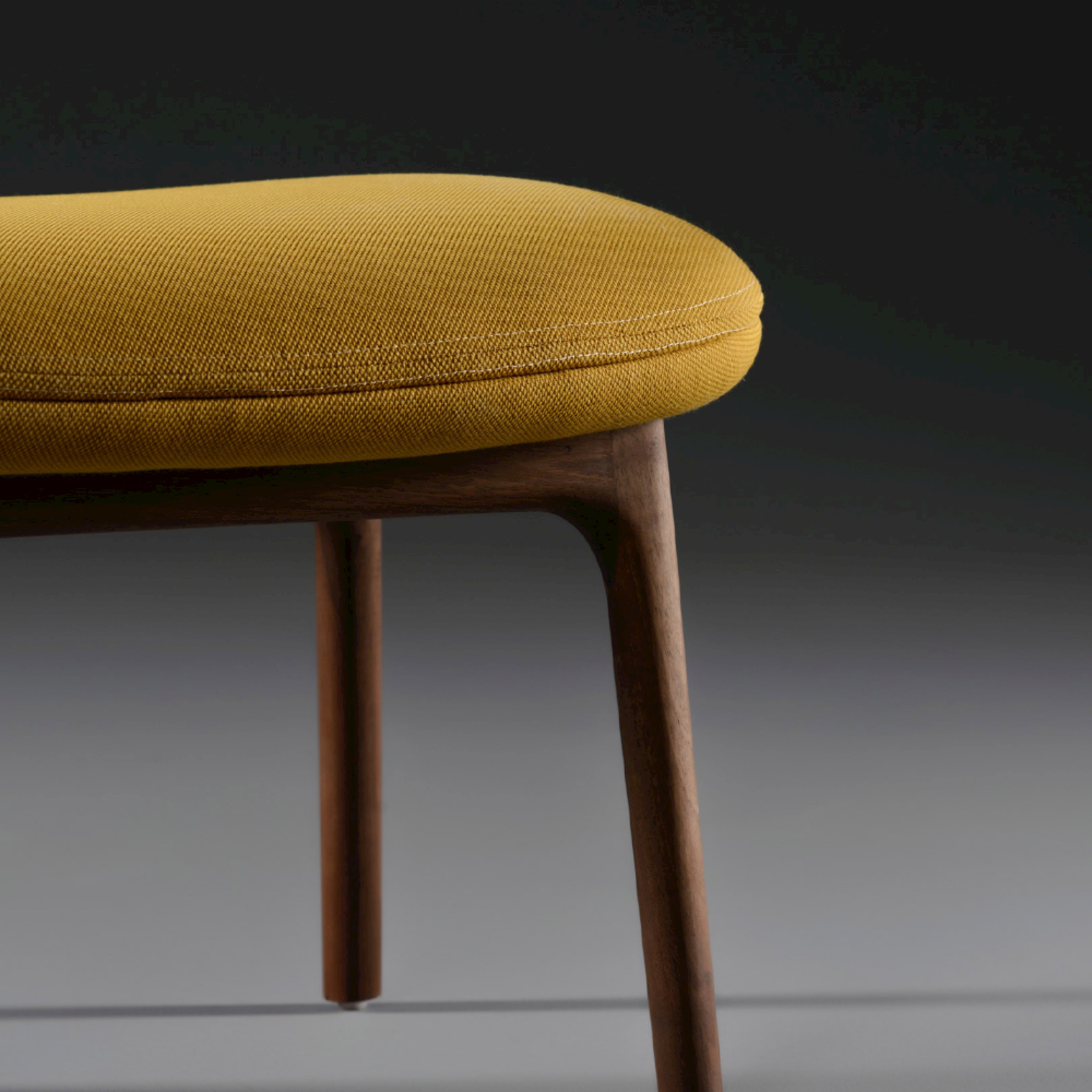 Close-up of the NEVA Ottoman by Artisan, featuring a mustard-yellow upholstered cushion and sleek, dark wooden legs, set against a dark gray gradient background. Its minimalist design elegantly captures modern Scandinavian lounge furniture aesthetics.