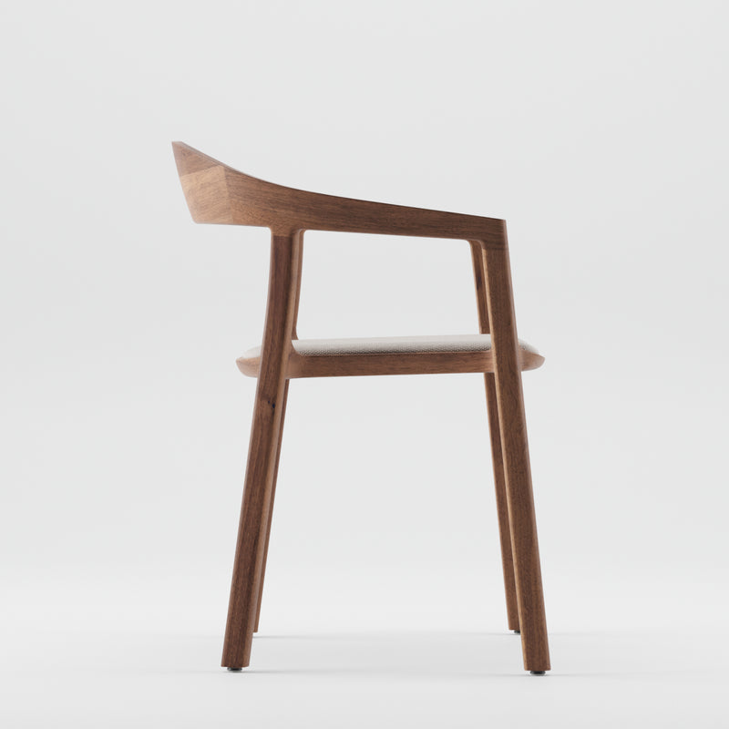 Side view of the TARA Armchair by Artisan, showcasing its minimalist design crafted from solid wood with a natural oil finish. It features a curved backrest, angled armrests, and a cushioned seat, standing on four straight legs against a plain white background.