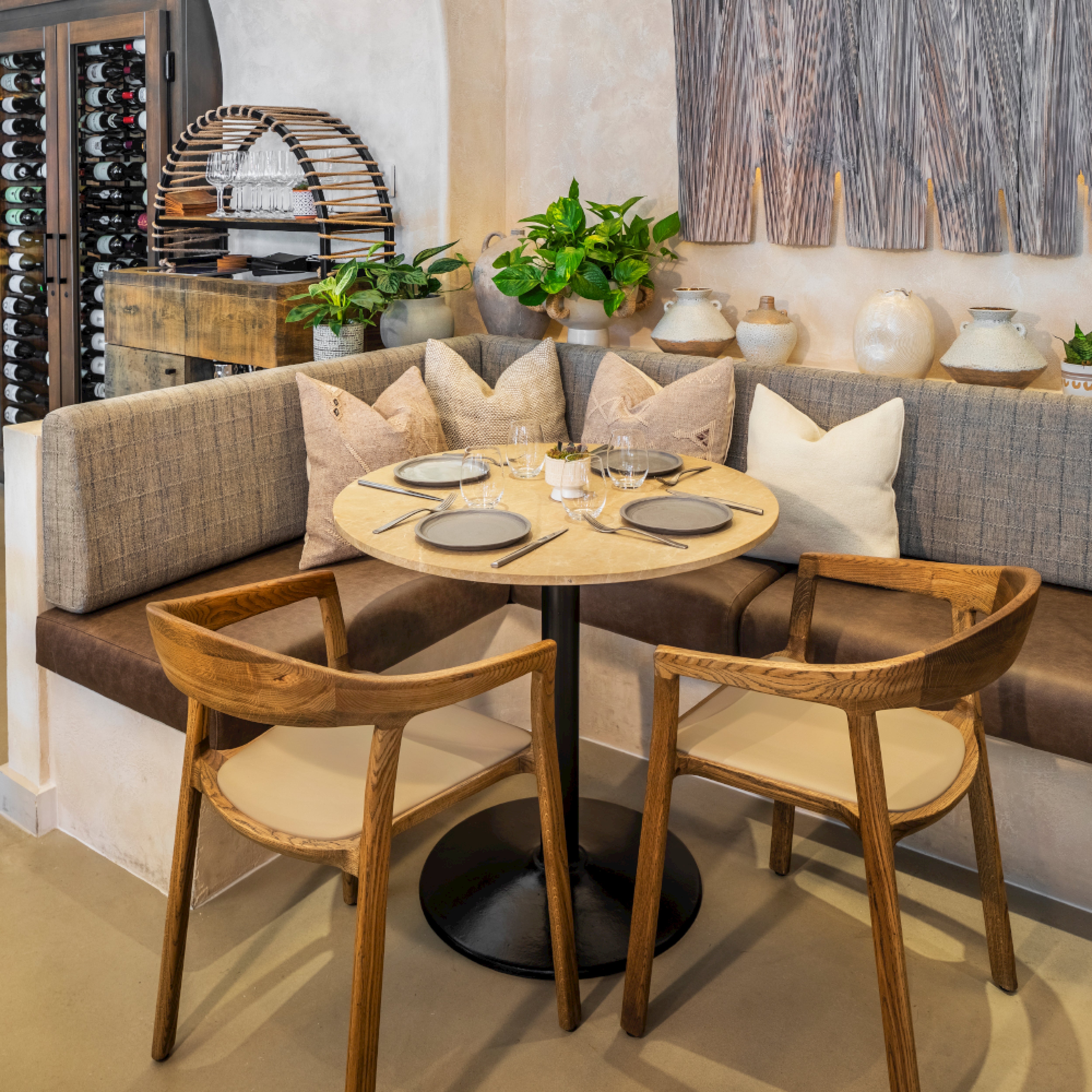 A cozy restaurant nook showcases a round table set for two with wine glasses and plates, surrounded by plush pillows. Two Artisan TARA Armchairs complement the seating. Nearby, wine bottles are stored with green plants and neutral decor made from eco-friendly materials to enhance the ambiance.