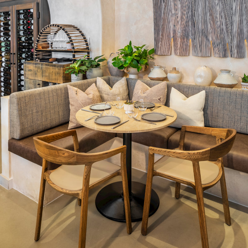 A cozy restaurant nook showcases a round table set for two with wine glasses and plates, surrounded by plush pillows. Two Artisan TARA Armchairs complement the seating. Nearby, wine bottles are stored with green plants and neutral decor made from eco-friendly materials to enhance the ambiance.
