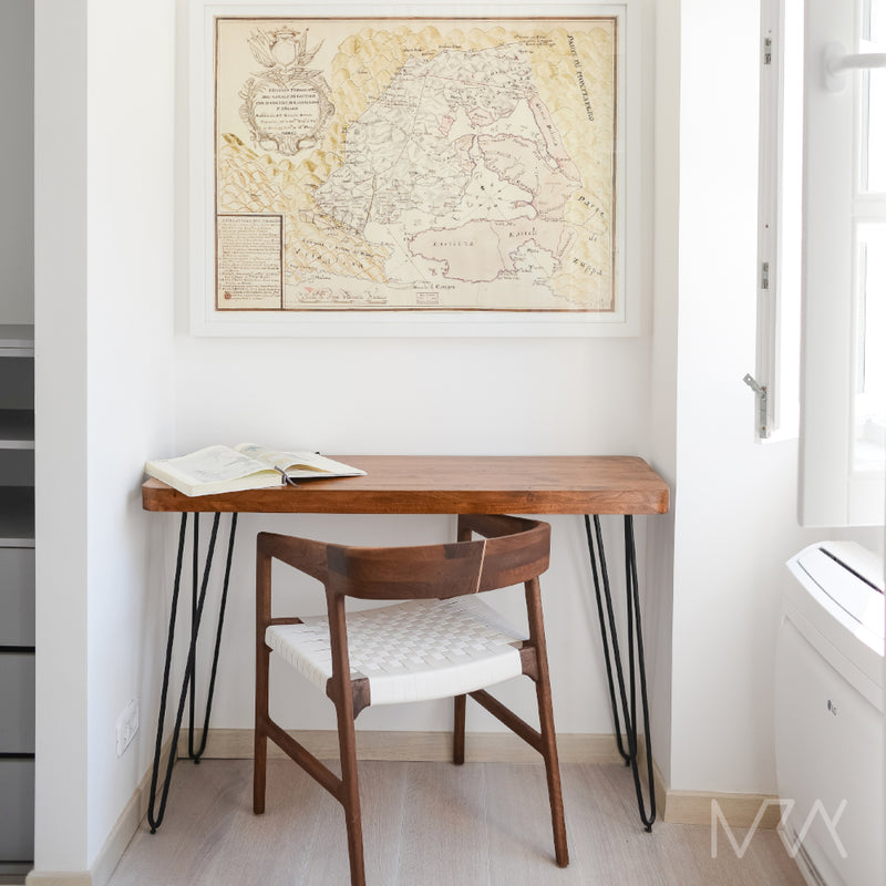 A minimalist study nook includes a wooden desk made from eco-friendly materials and the Artisan TESA Chair with a woven seat. An open book is on the desk, and a vintage map is framed on the wall, lit by natural light streaming through the nearby window.