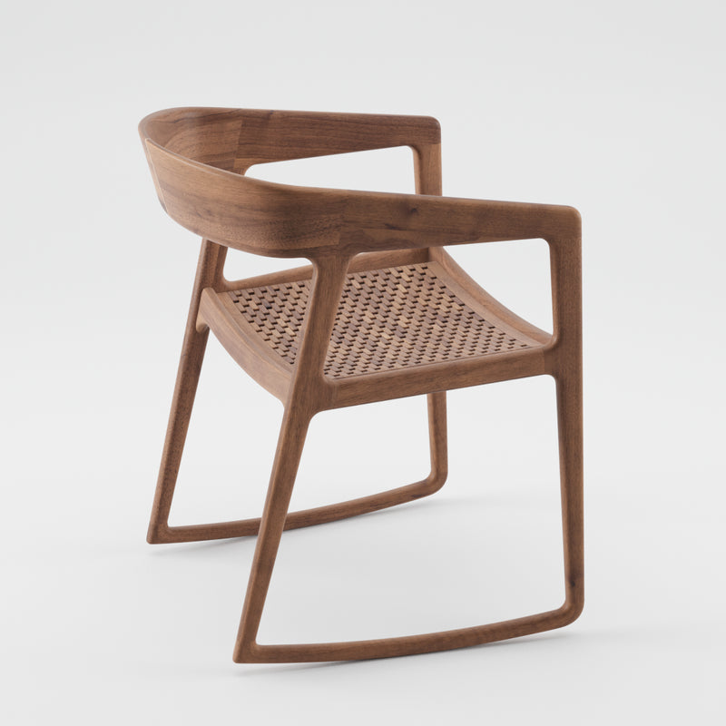 Featuring the Artisan brand, the TESA SWING CHAIR is a modern wooden chair with a side view. Made from solid wood and finished with natural oils, it boasts a curved backrest, woven seat texture, and sleek geometric armrests. Its eco-conscious style merges contemporary design with natural materials on a white background.