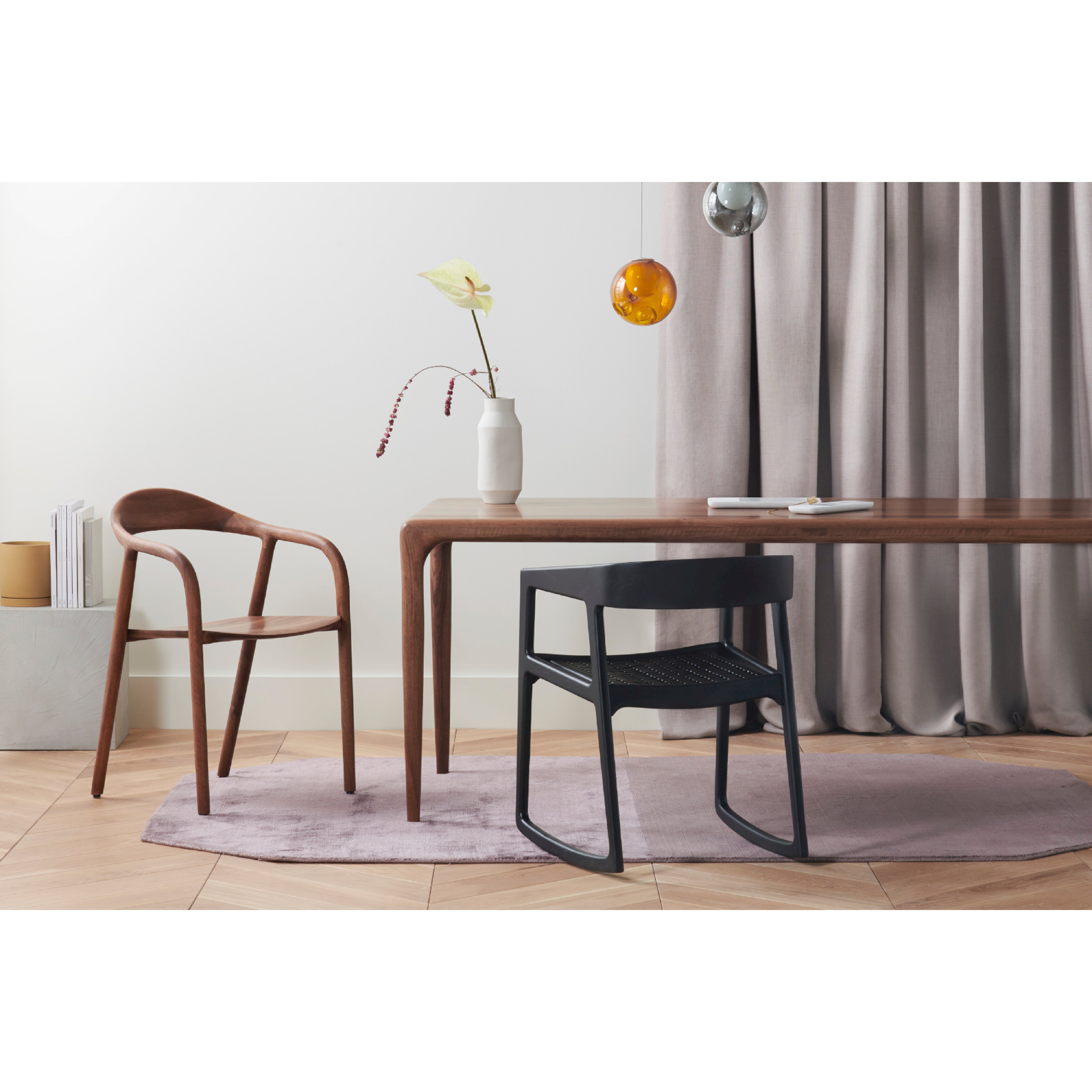 A modern dining room showcases a solid wood table, wooden chair, and the Artisan TESA SWING CHAIR on a lavender rug. A white vase with yellow and red flowers adorns the table. Two spherical pendant lights hang above, complemented by environmentally-friendly gray curtains in the background.