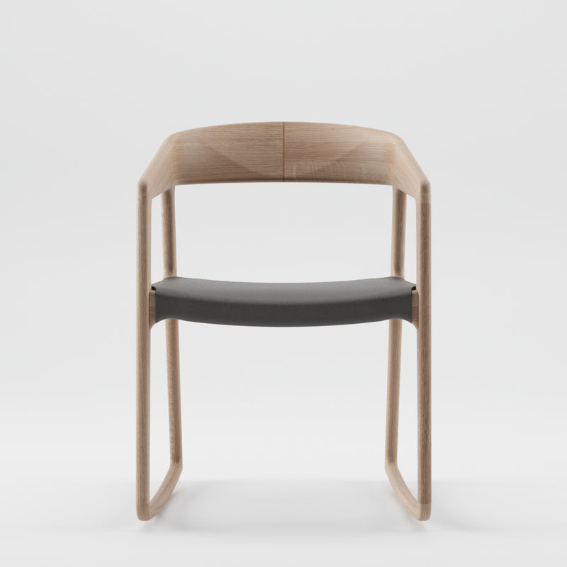 The Artisan TESA SWING CHAIR features a minimalist design with a curved backrest, crafted from solid wood and finished with natural oils. It has a dark cushioned seat, smooth edges, and an open back, set against a plain white background for an eco-friendly touch.