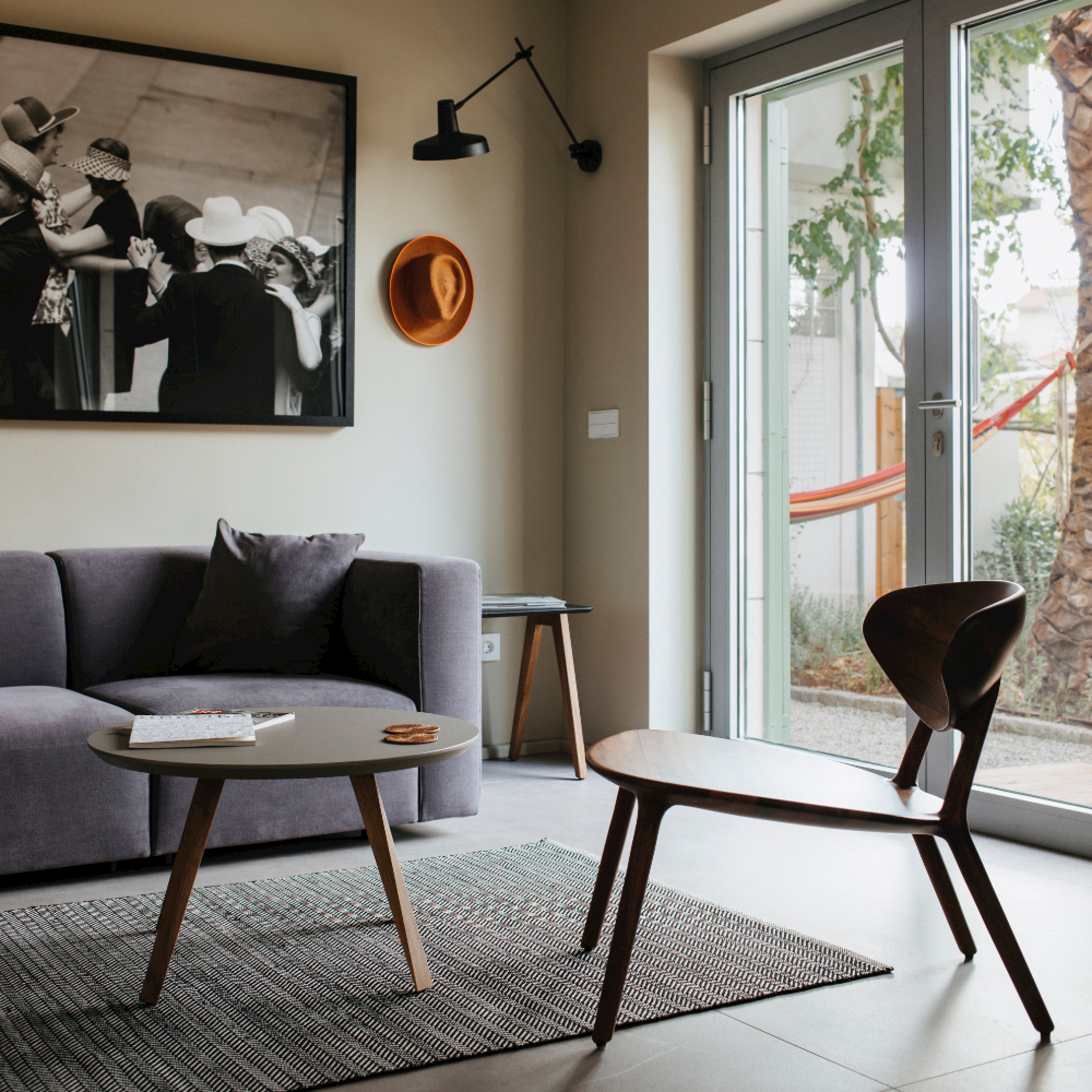 A modern living room features a gray sofa, round coffee table, and the Artisan WU Lounge Chair crafted from solid wood. A black-and-white photo and an orange hat adorn the wall. Large windows reveal a hammock outside, while a rug partially covers the tiled floor with eco-friendly materials.
