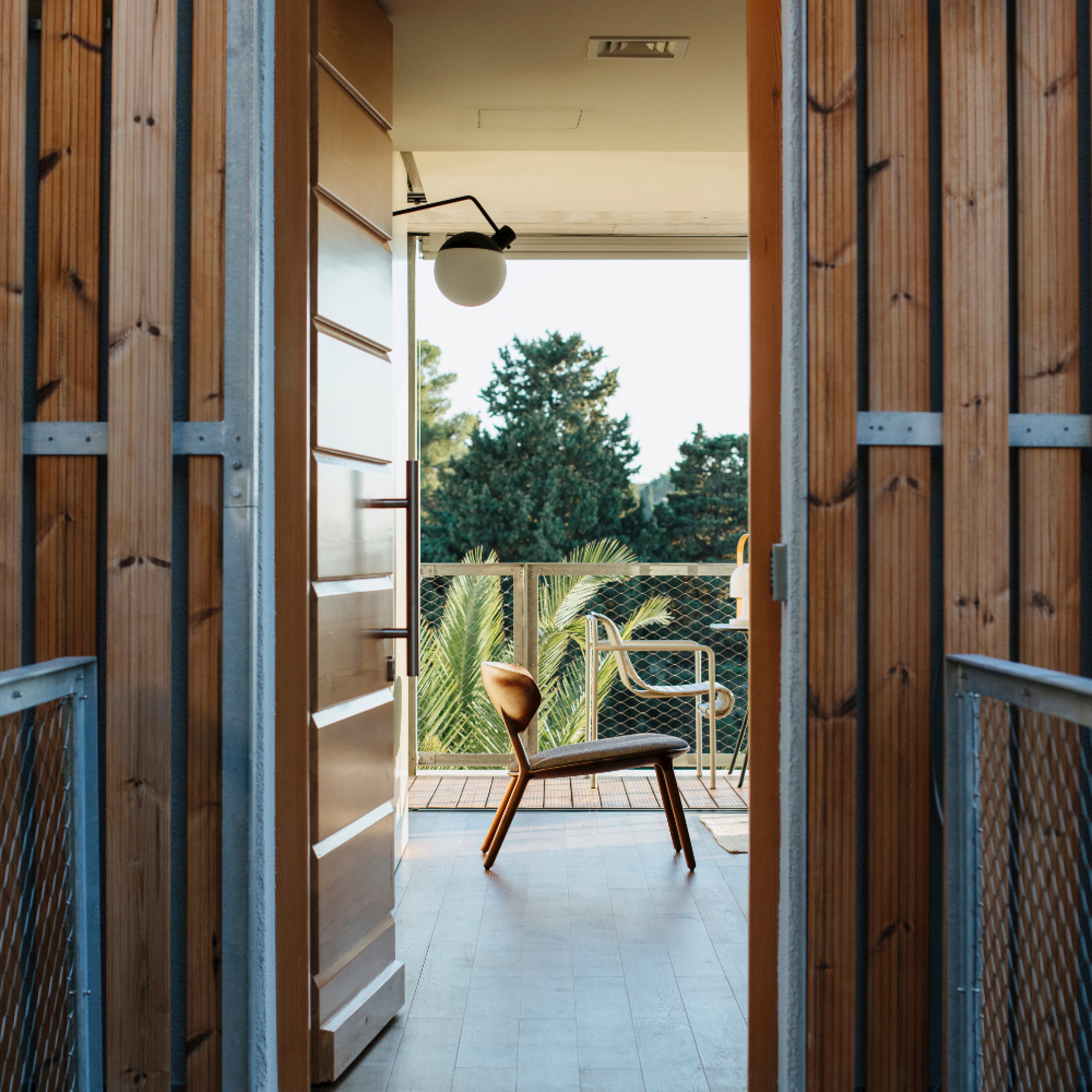 The WU Lounge Chair by Artisan, crafted from solid wood and treated with natural oils, is placed on a balcony. Framed by open wooden doors against lush green trees and a clear sky, it creates an eco-friendly and serene outdoor setting.